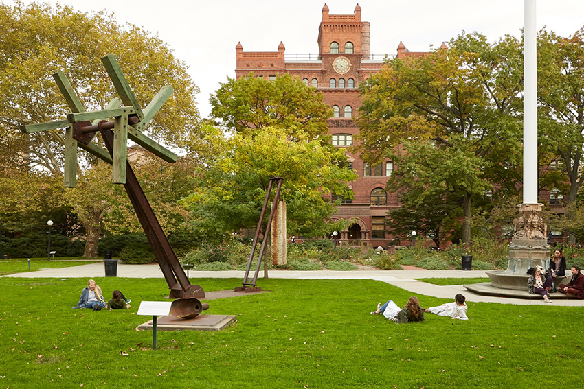 Students lay on a lawn with scattered metal statues and marble fountain. There is a red brick building in the background.
