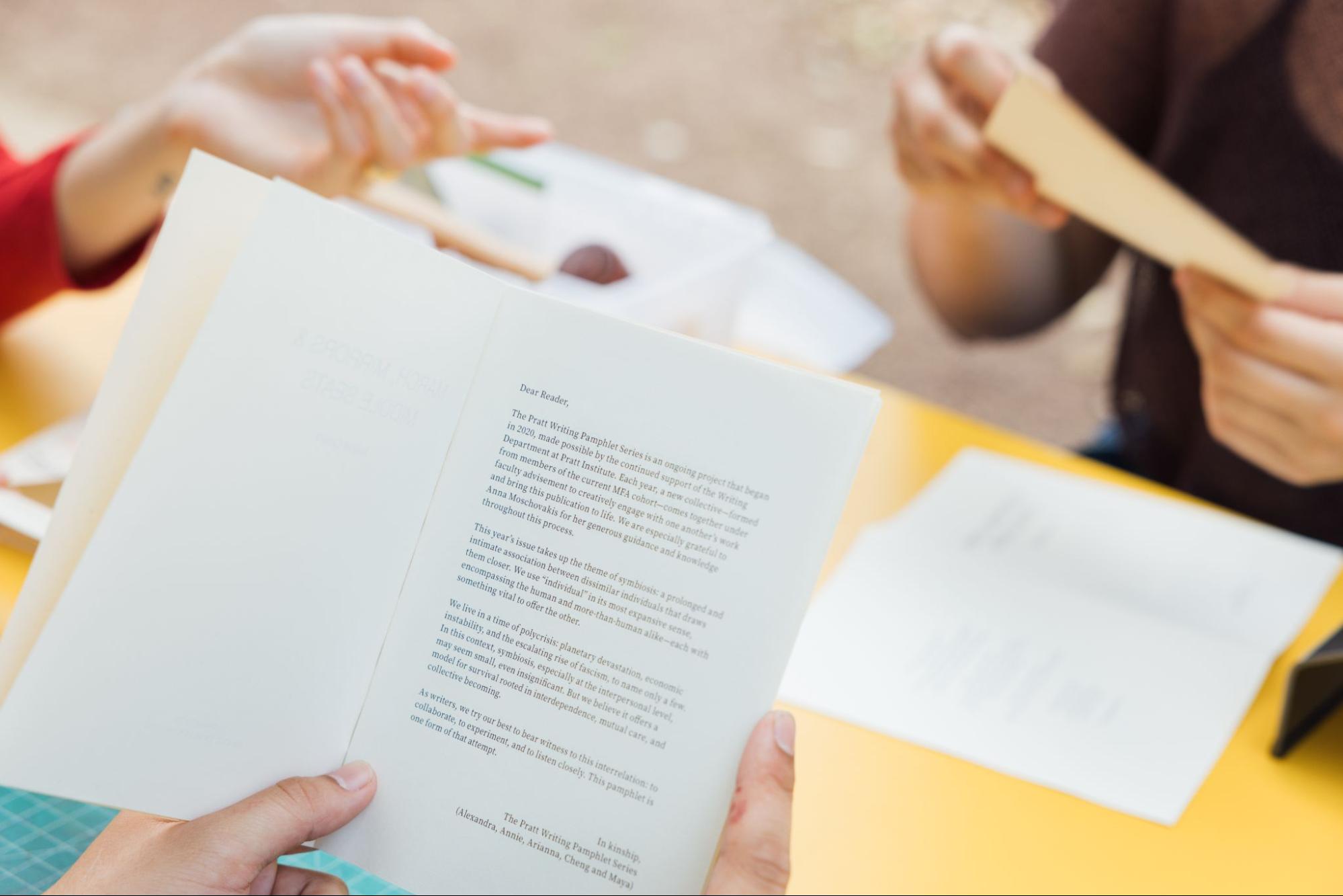 Close-up view of hands holding an open booklet over a yellow table surface. The visible page includes a printed letter beginning with “Dear Reader,” formatted in paragraph text. In the blurred background, another person holds a book, suggesting a shared reading or discussion setting.