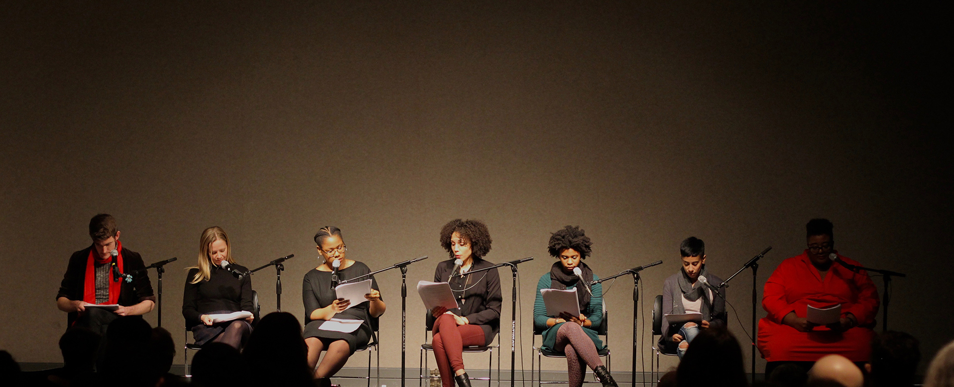 A diverse group of people sit on stage as they each read from papers they are holding in their hands. They all have microphones in front of them.