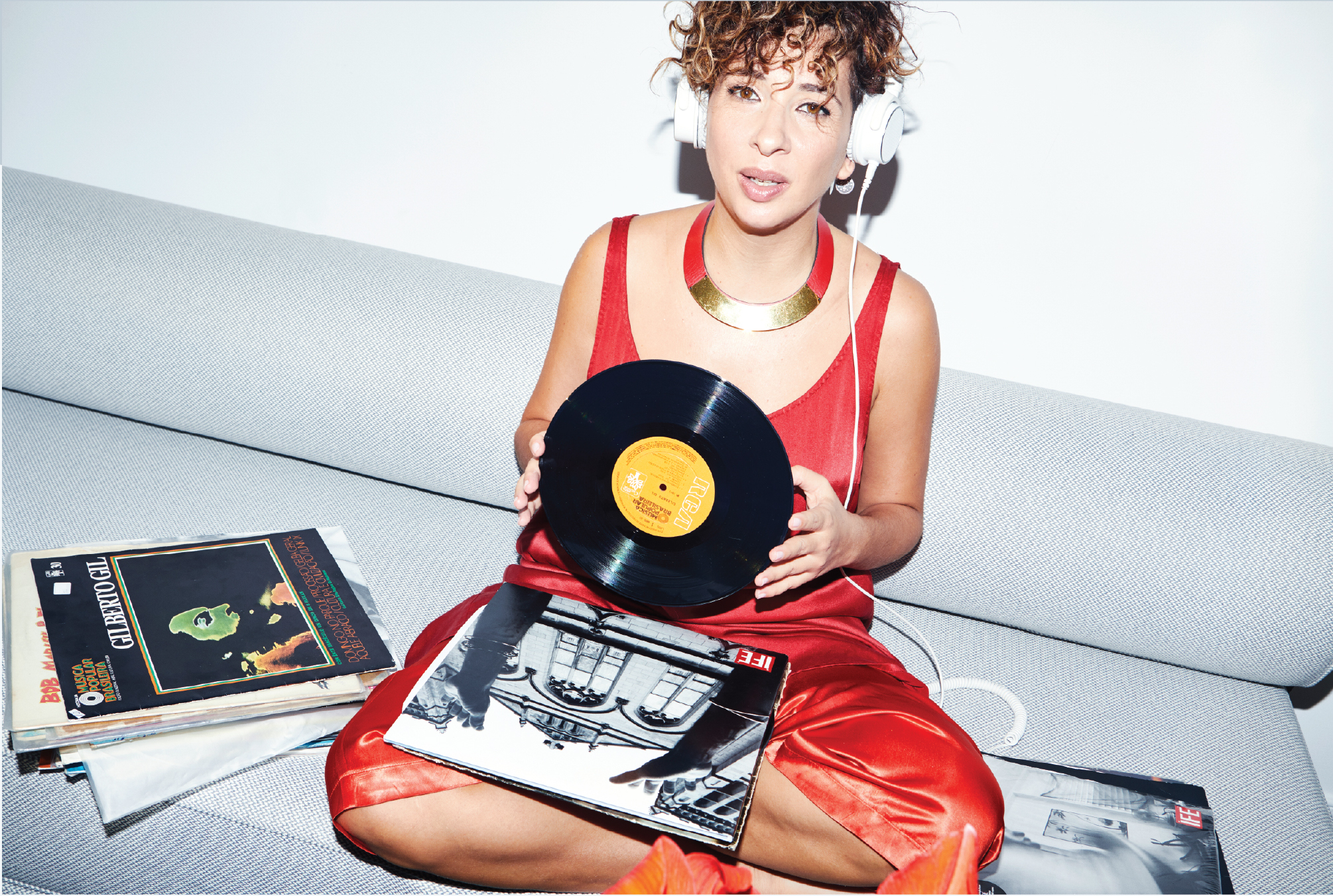 A woman with short, curly brown hair sits cross-legged on a light gray couch against a plain white wall. She wears a red satin dress with thin straps, a bold gold necklace, and white over-ear headphones. She holds a black vinyl record with a yellow RCA label in both hands, looking directly at the camera with a confident expression. Around her are several vinyl records and album covers, including one prominently featuring an architectural image and another labeled 