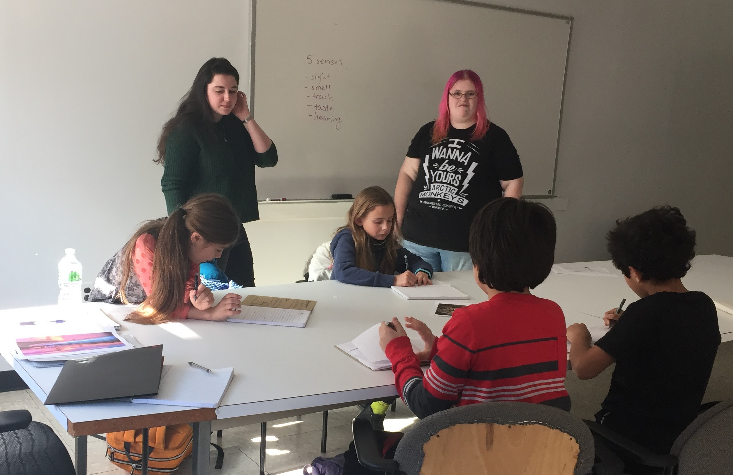 Two adult facilitators—one with long dark hair and the other with bright pink hair—stand at the front of a classroom near a whiteboard with the words 