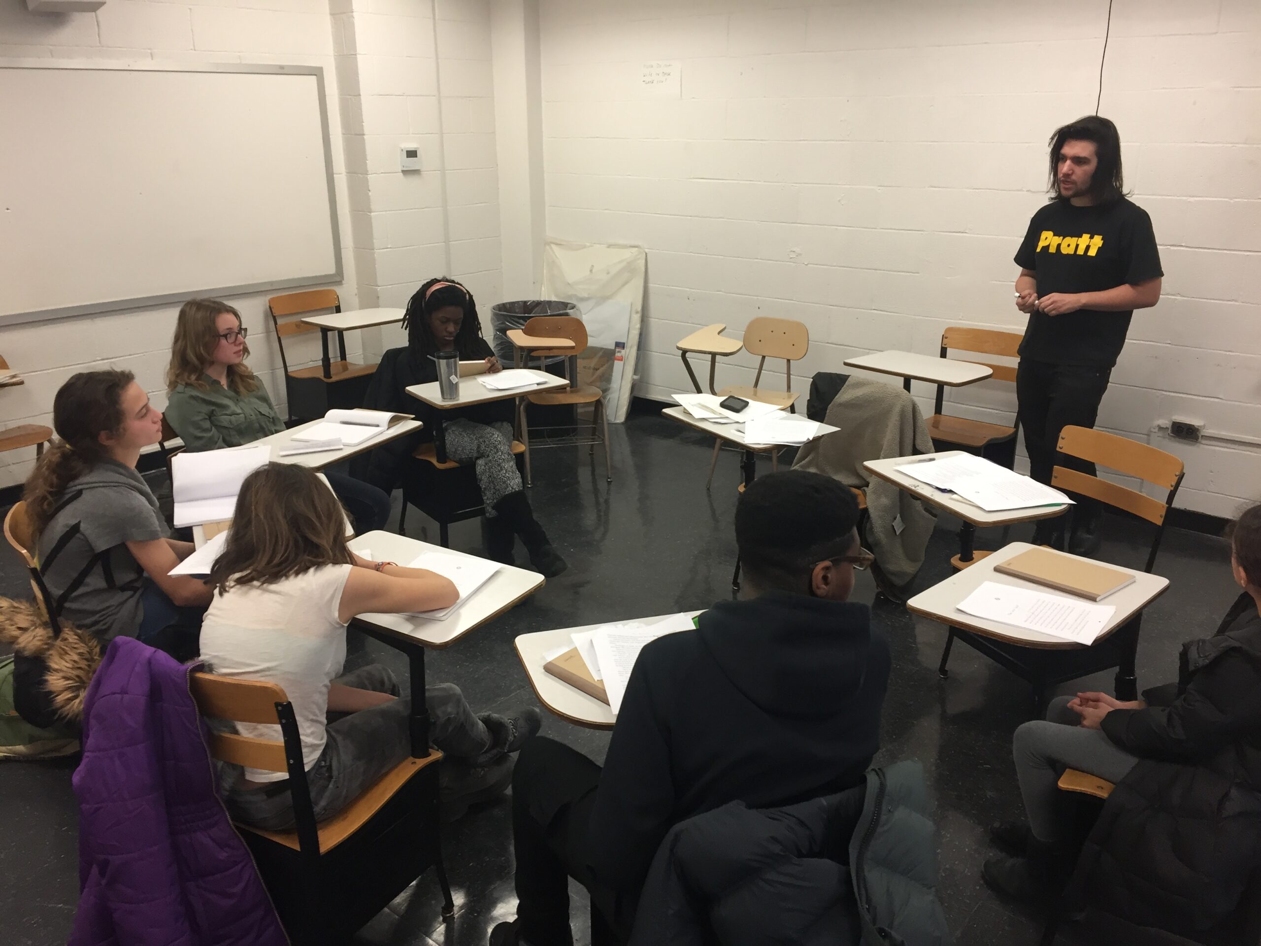 A classroom setting with a male instructor in a black Pratt t-shirt standing and addressing a semi-circle of seven students, all seated at separate desks with notebooks and papers. The room has white cinderblock walls, black flooring, and a blank whiteboard in the background.