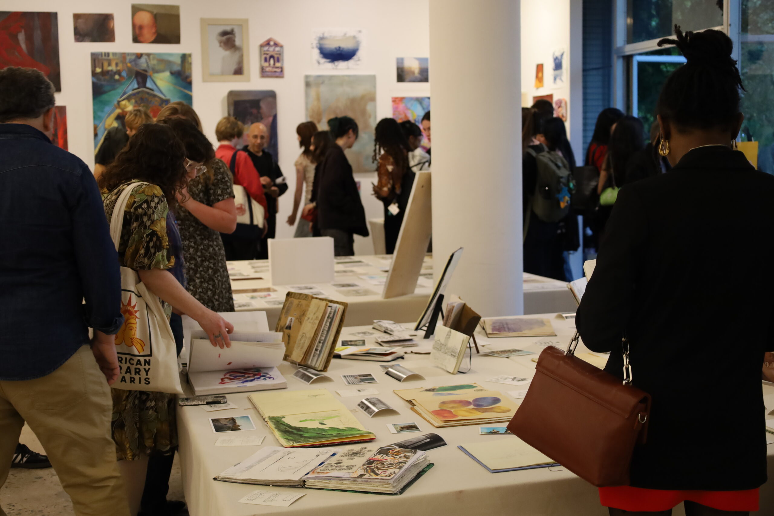 A group of people is attending an art exhibition in a bright, spacious gallery. Various artworks hang on the walls, while tables covered with documents, sketches, and art supplies occupy the center. A woman in a floral dress examines a booklet, while others engage in conversation and observe the displayed pieces. A column stands in the middle of the room, and large windows allow natural light to fill the space.