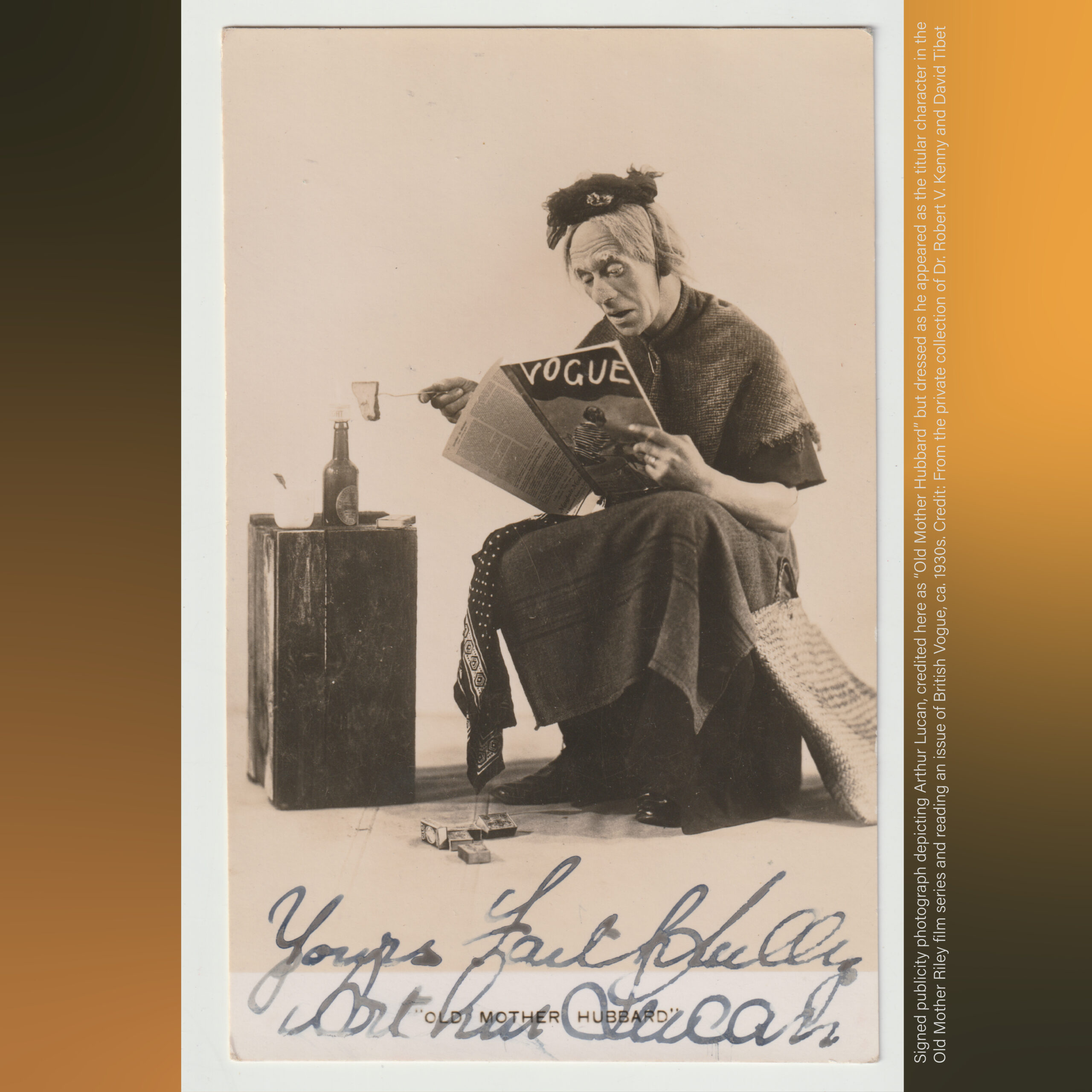Sepia-toned vintage photograph of a costumed performer seated on a low stool, wearing layered clothing and a head covering, holding and reading a copy of Vogue magazine. One hand extends outward holding a small object above a bottle placed on a wooden crate beside them. Small boxes lie on the floor near their feet. The image includes a handwritten signature across the bottom margin. Along the right edge, vertical printed text provides contextual and credit information about the photograph and its historical background.