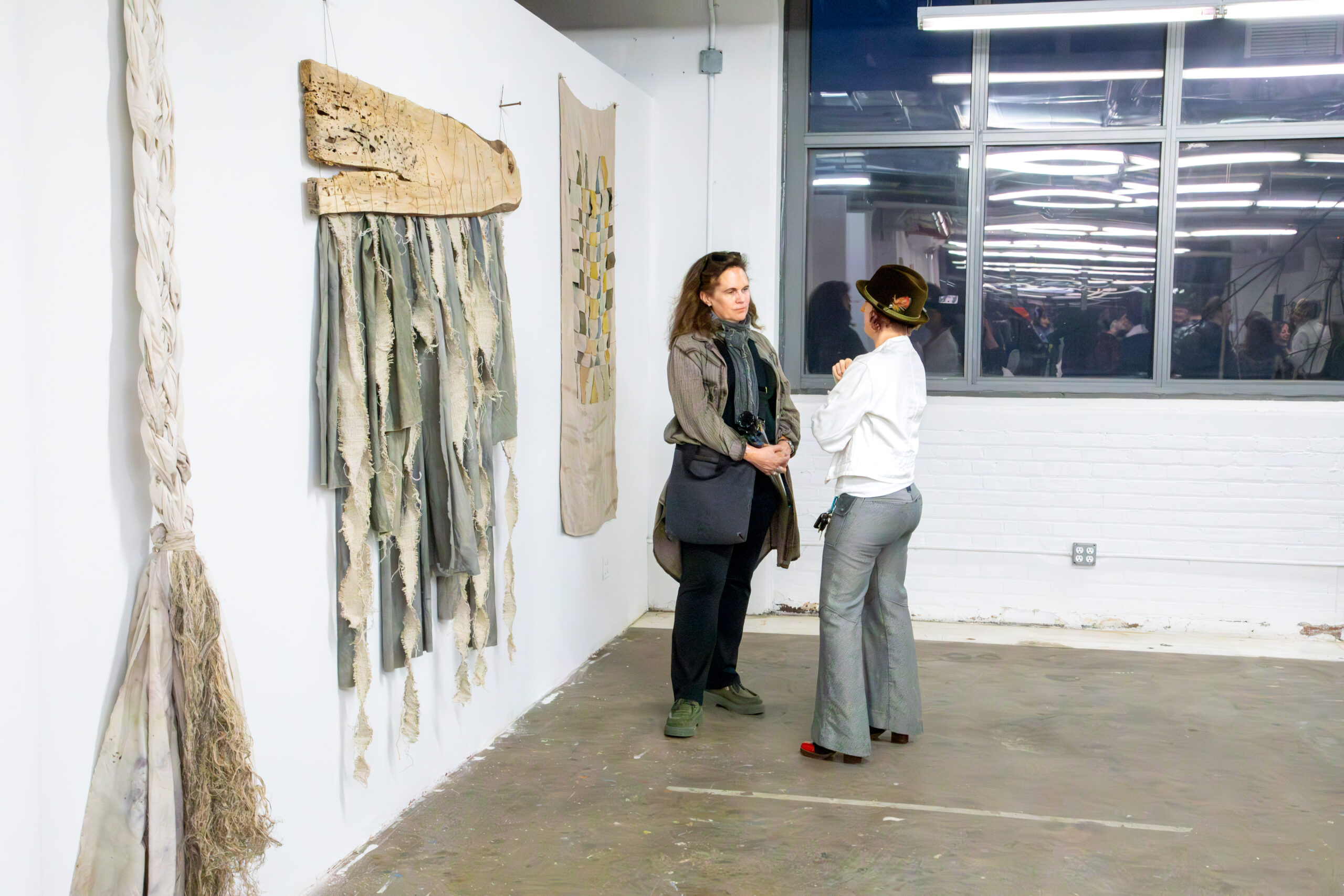 Two people stand facing each other in conversation inside a white-walled gallery space. Textile-based artworks hang vertically on the wall to the left, including braided and layered fabric pieces with frayed edges. A large window on the right reflects ceiling lights and a crowd gathered in an adjacent space. The floor is concrete, and the setting appears to be an exhibition opening or gallery event.
