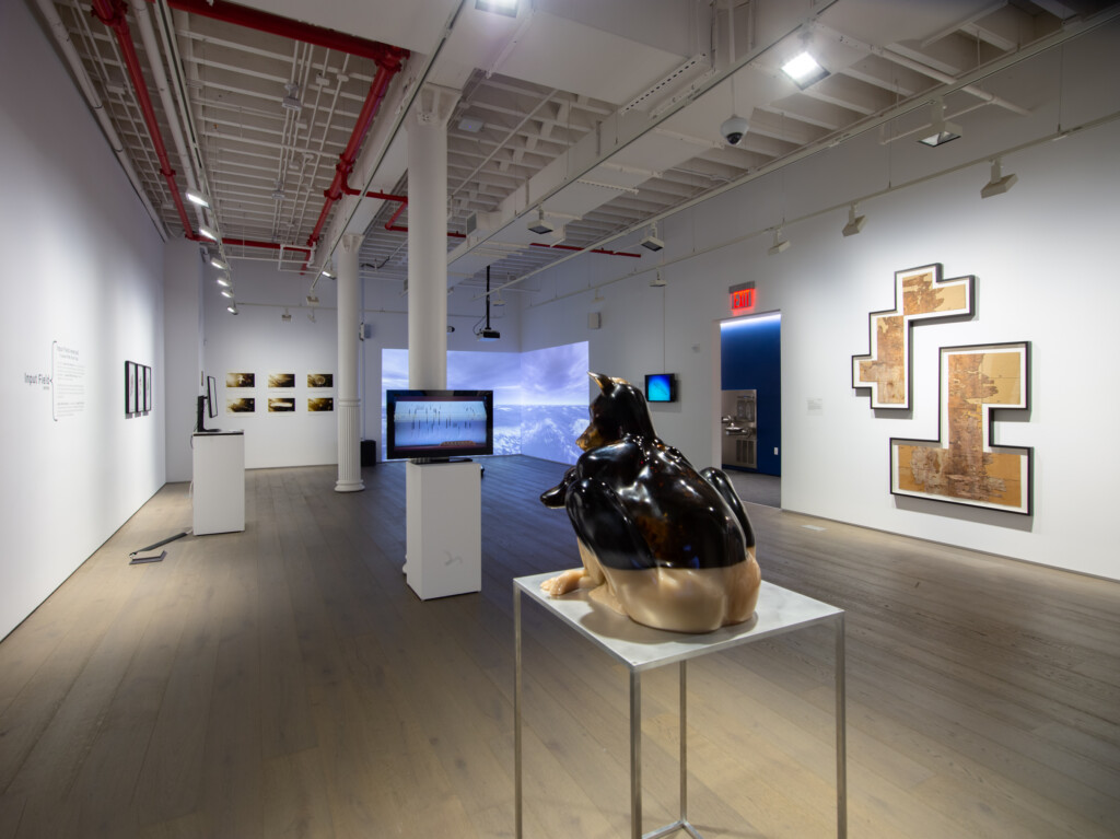 a large gallery room, with artworks that include paintings, video monitors, sculptures, and installations, with white walls and a wooden floor