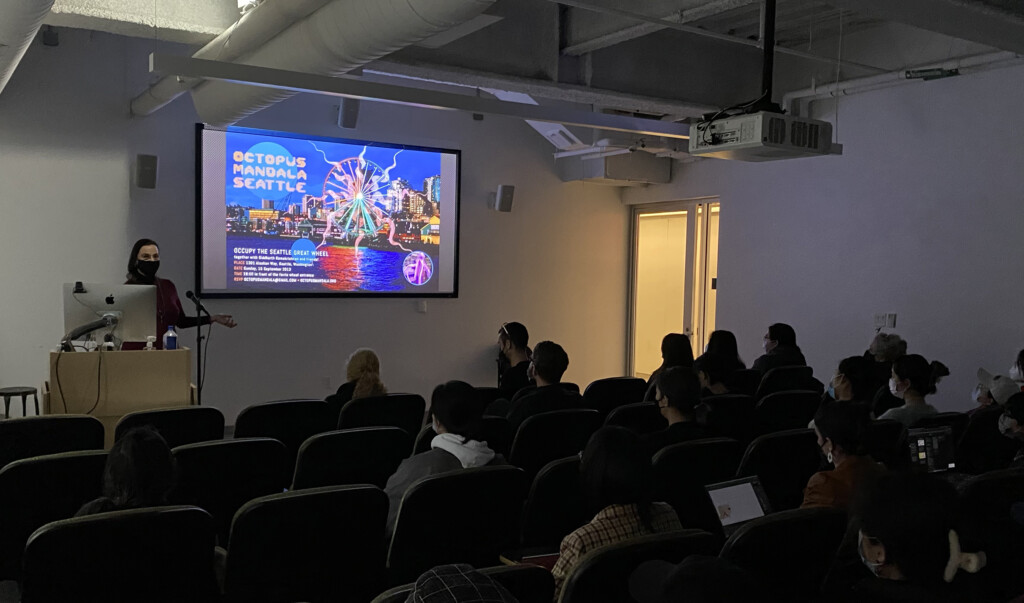 Auditorium with people seated, watching a presenter at a podium, and a projection screen displaying information