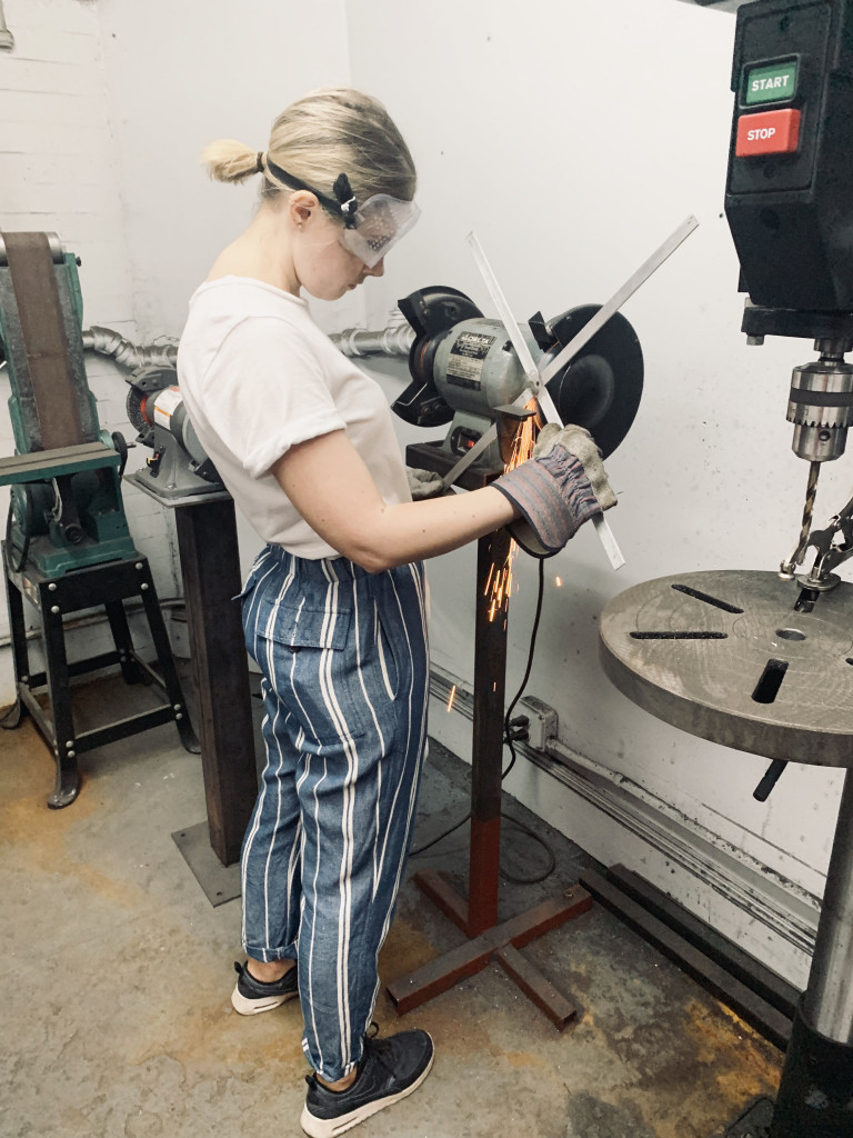 a woman statnding at a buffer for metal objects, taking imperfections from what she is holding, in a workshop, wearing safety goggles