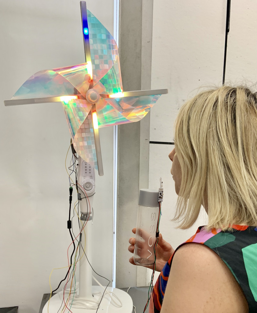 A blonde-haired woman looking at the model of a windmill, with iridescent foil material on the outside of the windmill's rudders, which have lightbars attached to them