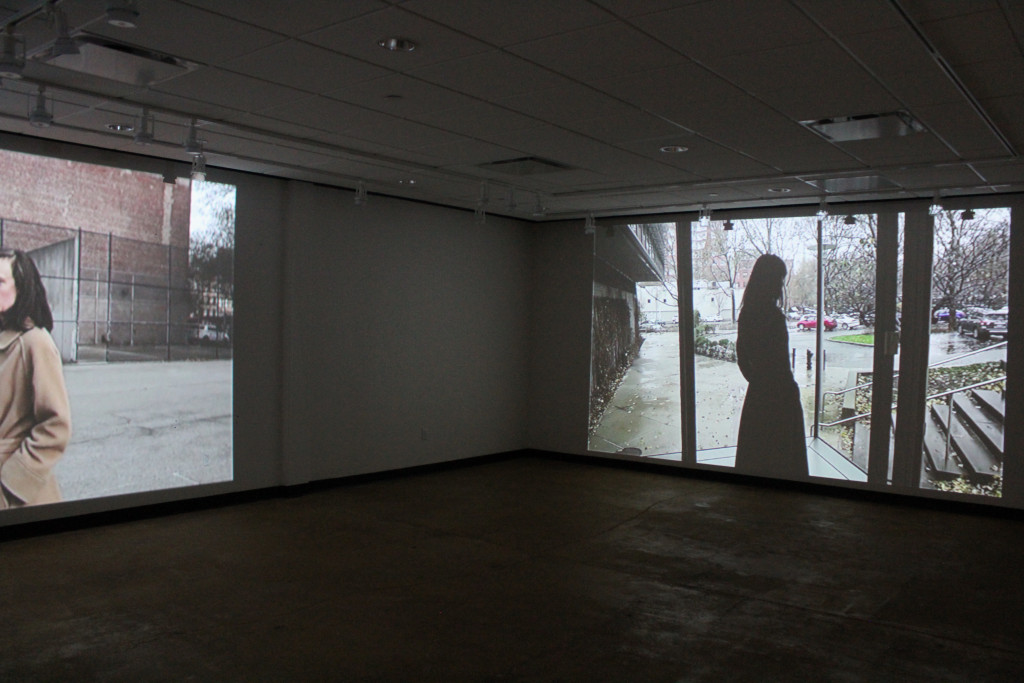 dark room with two movie projections, one with woman walking in urban plaza courtyard, another with a woman standing in a building, looking down at the courtyard