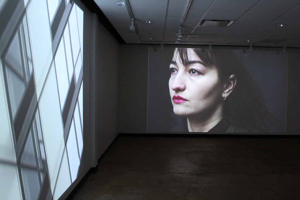 dark room with two movie projections, one with building windows and another with a womans face looking upward