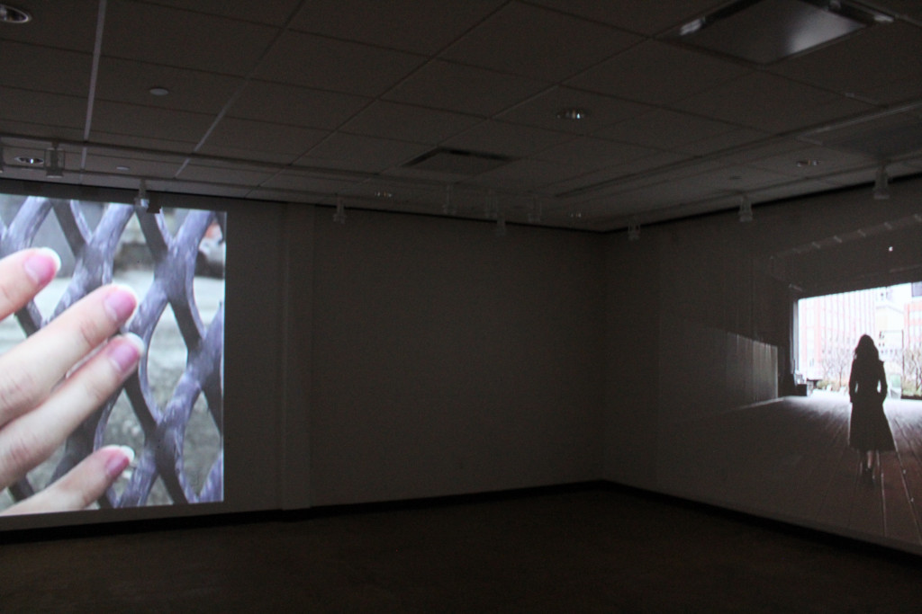 dark room with movie projection, depicting hand with long nails and on a separate wall, a projection of a woman walking through a dark train depot