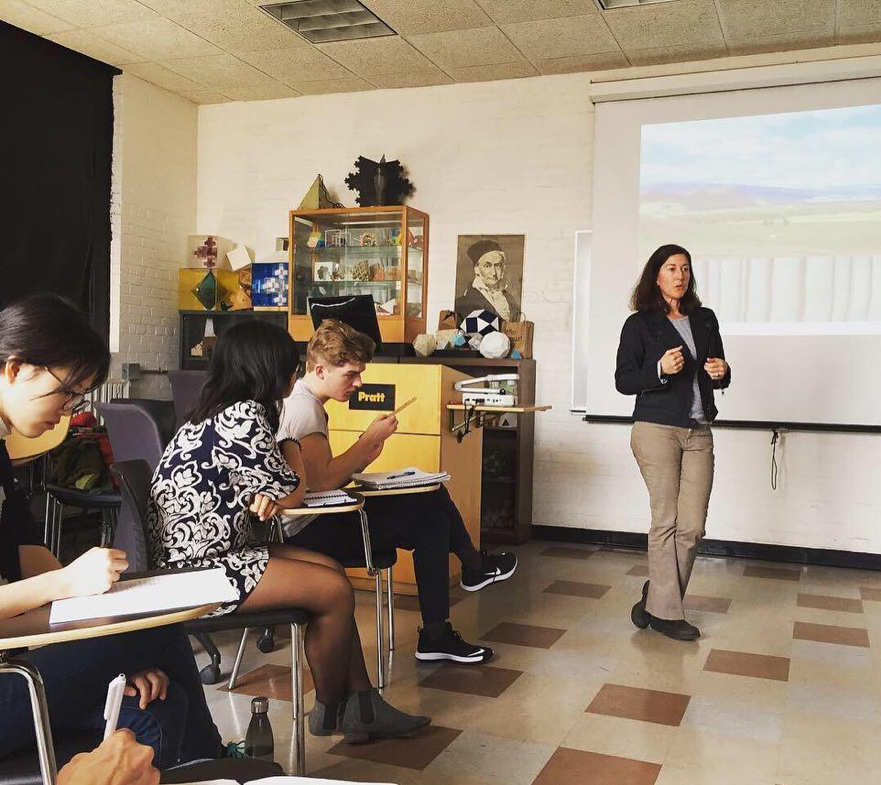 students watching teacher speak, in a classroom, in front of a projector
