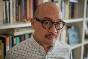 A man with a shaved head, mustache, and tortoiseshell round glasses stands in front of a book-filled shelf. He wears a white and green plaid button-up shirt and gazes calmly at the camera. The background features books and soft lighting, suggesting a study or personal library setting.