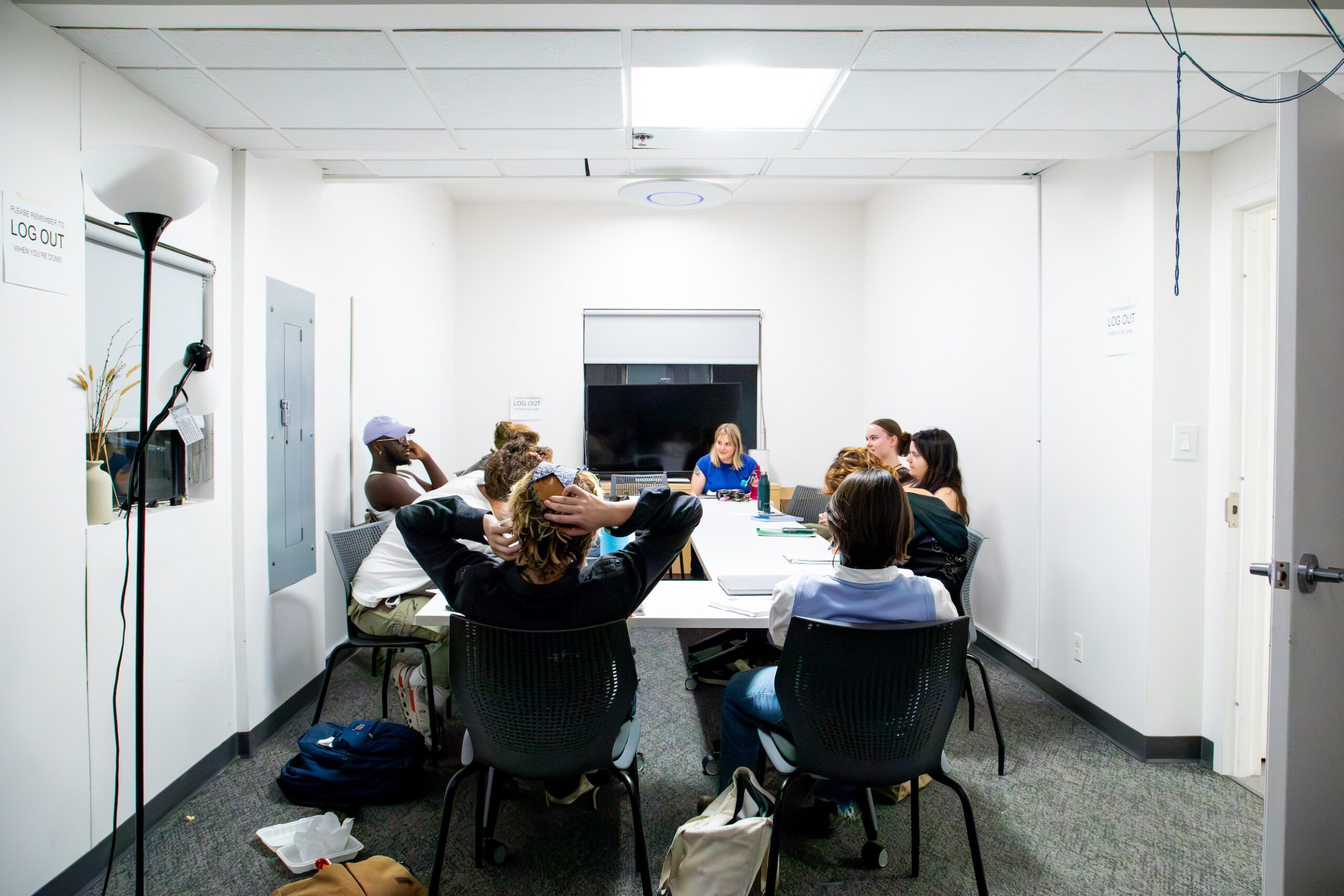 A group of students sits around a white conference table in a small, well-lit classroom. They are engaged in a discussion led by a person seated at the far end of the table. The students appear focused and attentive, with notebooks, drinks, and personal belongings scattered on the table.