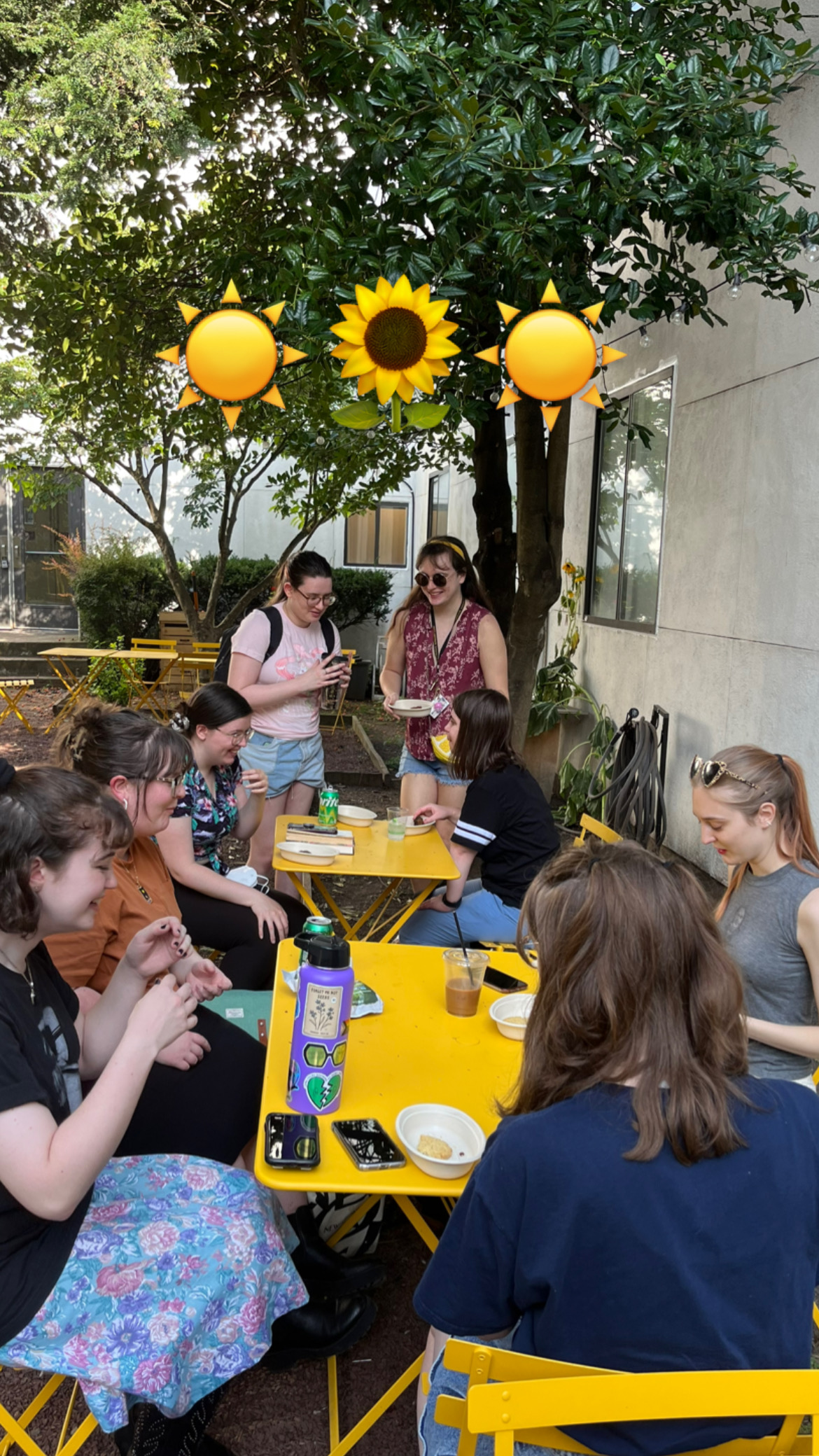 A group of students gathers at yellow tables in a shaded outdoor area, enjoying snacks and drinks. Some are standing and chatting while others are seated. Emojis of two suns and a sunflower are overlaid at the top of the image.