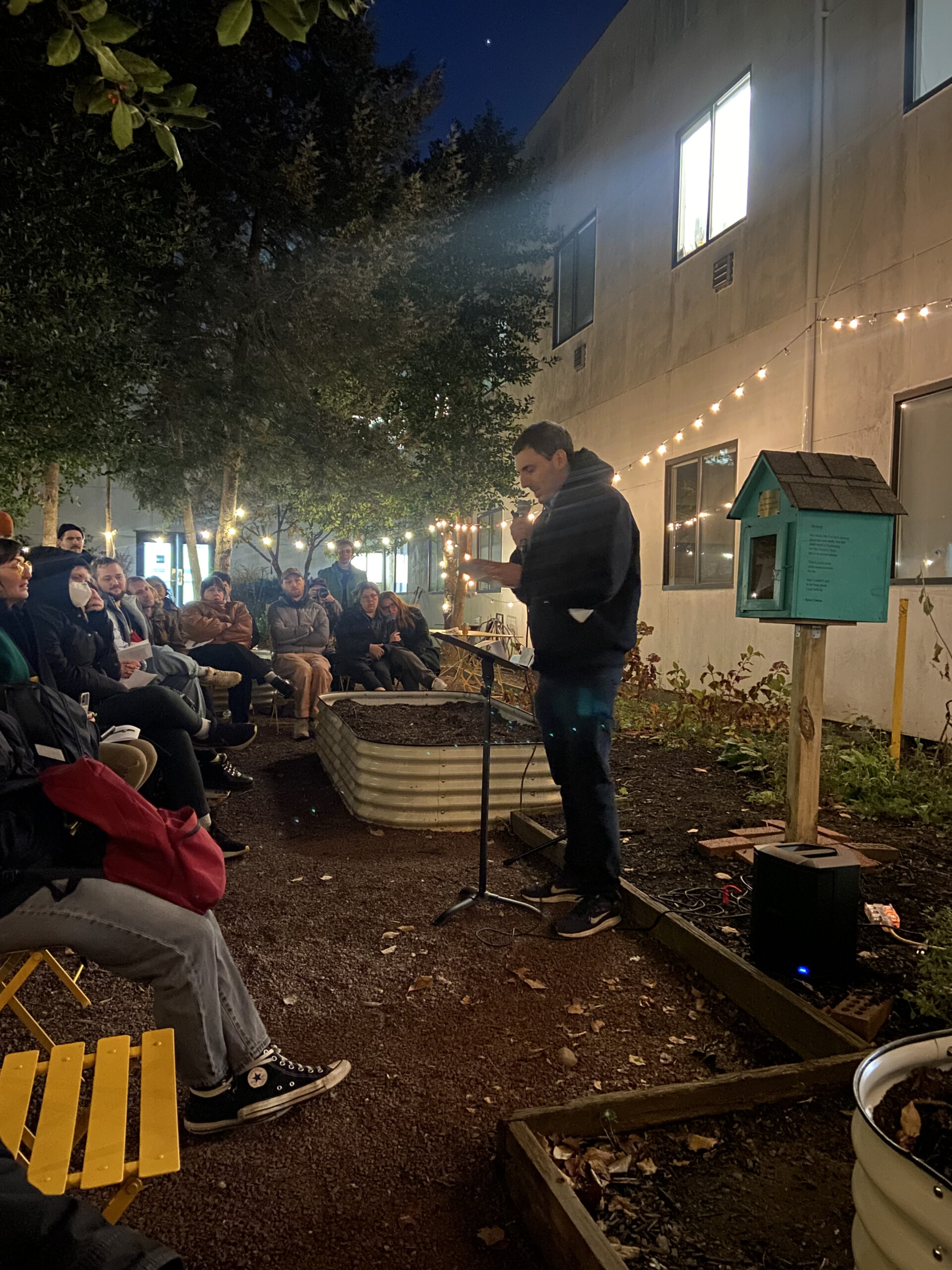 At night, a man reads from a paper at a microphone in an outdoor garden setting, illuminated by string lights. The audience is seated on yellow chairs facing him, and a small turquoise library box stands nearby, marking the event space.