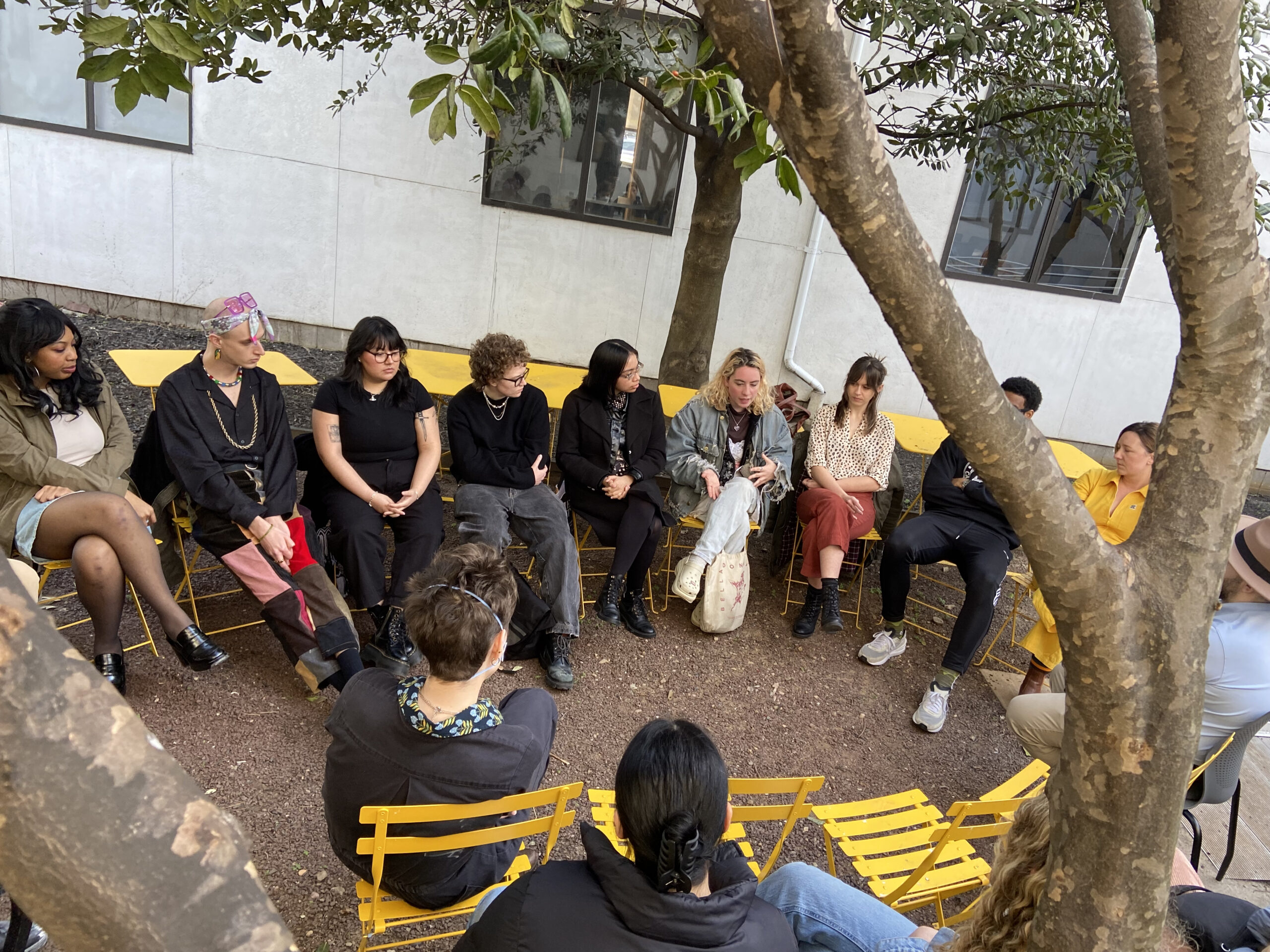 A diverse group of people sits in a circle of yellow chairs in an outdoor courtyard, engaged in discussion. The setting is casual and shaded by trees, with a white building wall in the background. Several participants gesture as they speak, suggesting an open dialogue or community conversation.