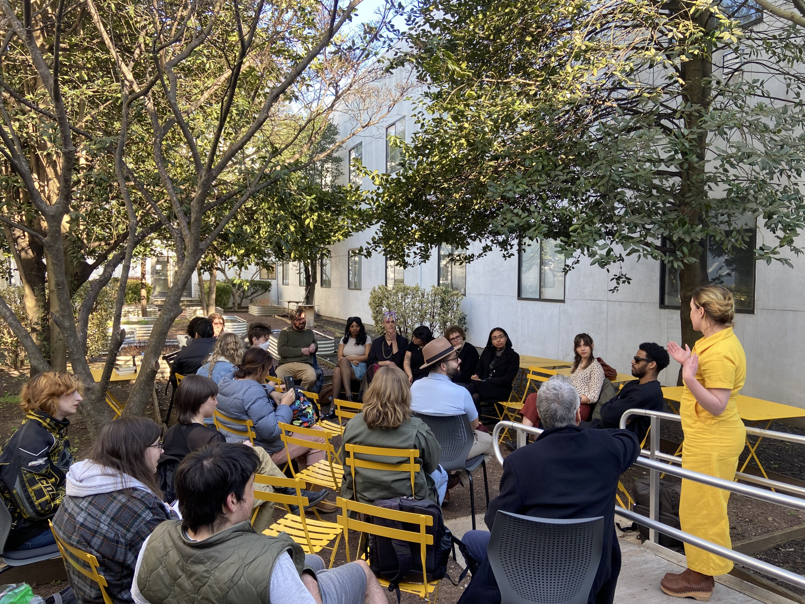 An outdoor roundtable event takes place in a garden courtyard. A woman in a yellow jumpsuit speaks to a seated group of students and alumni on yellow chairs. The event is framed by trees, a white building, and ambient daylight.