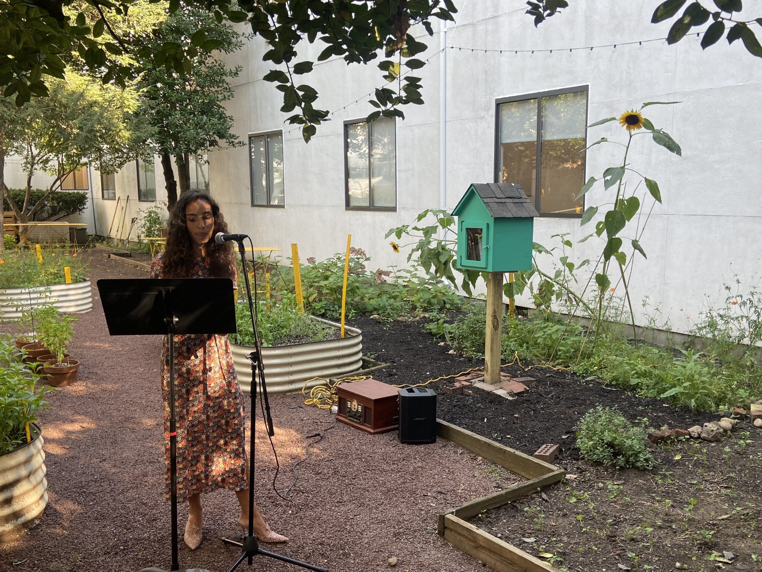 A woman in a floral dress stands at a microphone, reading aloud in a garden setting. She is positioned beside a small green library box, surrounded by sunflowers and planter beds. The event takes place in daylight, beneath leafy trees.
