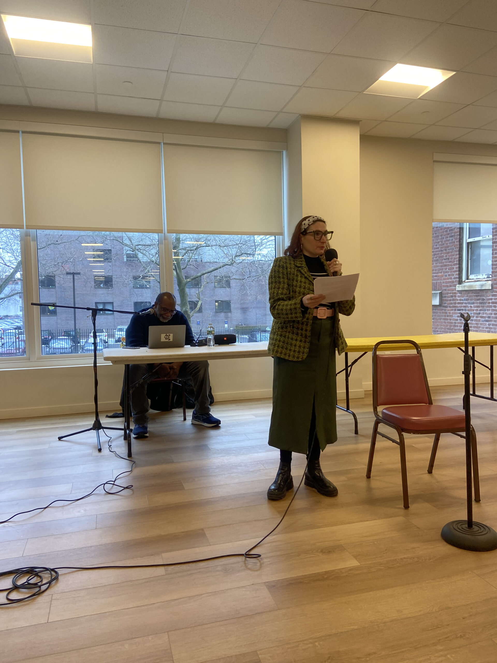 A woman in a green checkered jacket and long green skirt speaks into a microphone while reading from a page. She stands in a well-lit room with large windows. Behind her, Fred Moten sits at a table with a laptop, preparing to present.