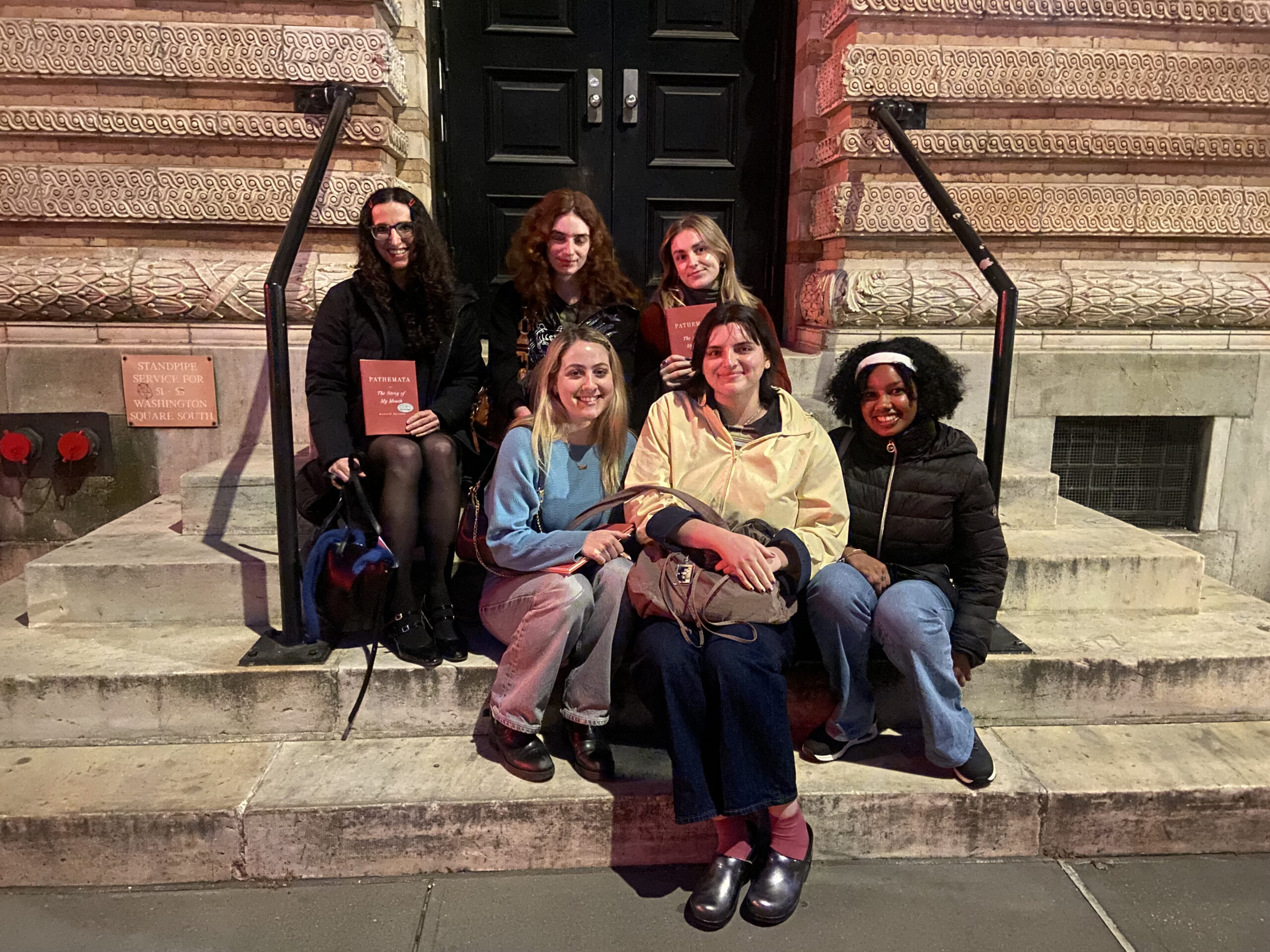 Six women sit on the stone steps of a historic building at night, holding red books titled *Patremata*. They smile and pose for the photo under warm lighting, with detailed architectural carvings and black double doors behind them. A stone plaque reads