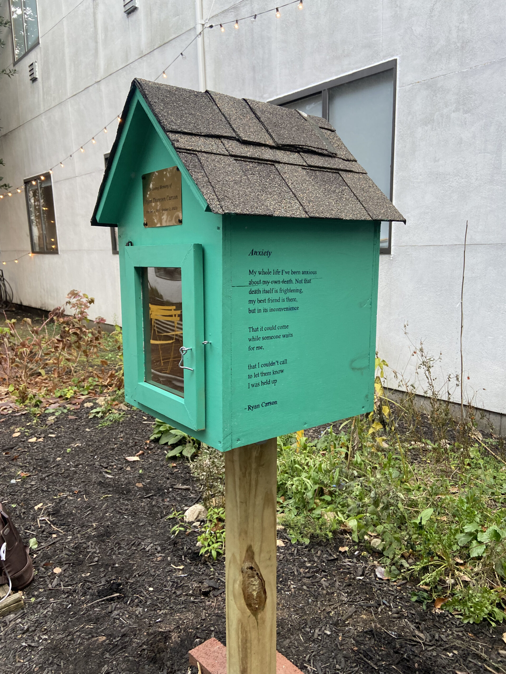 A small turquoise wooden library box with a dark shingled roof stands in a garden. A poem titled