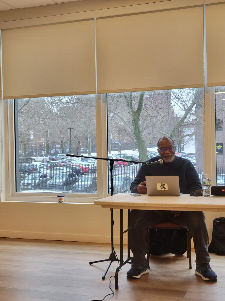 Fred Moten sits behind a table with a laptop, speaking into a microphone during a memorial reading event. The room is softly lit, with large windows showing a view of parked cars and bare trees outside. A water bottle and papers rest on the table.