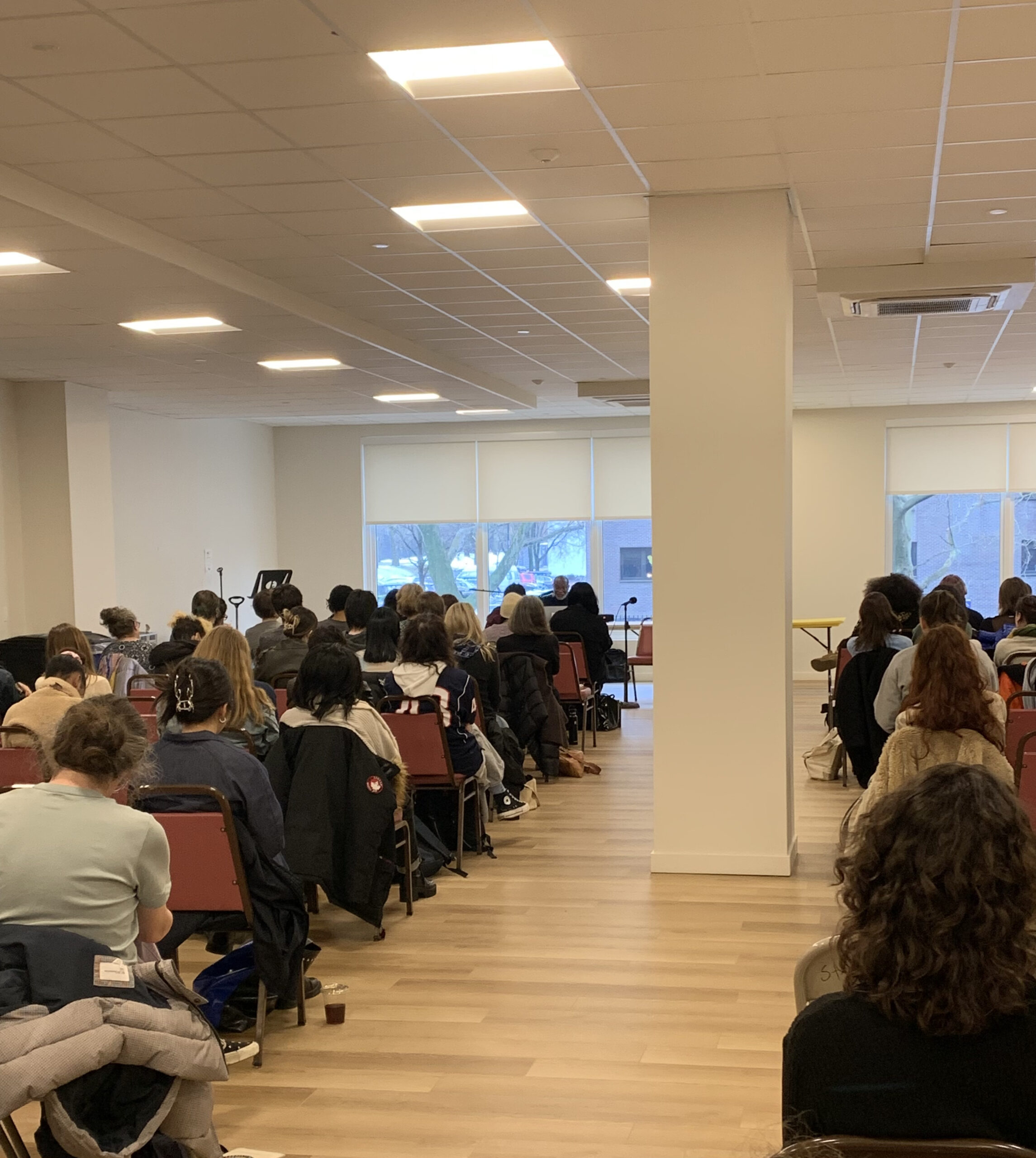 A wide view of a reading event in a brightly lit room. Attendees fill rows of chairs facing a front area with microphones and a laptop. Large windows in the background show trees and a parking lot outside.