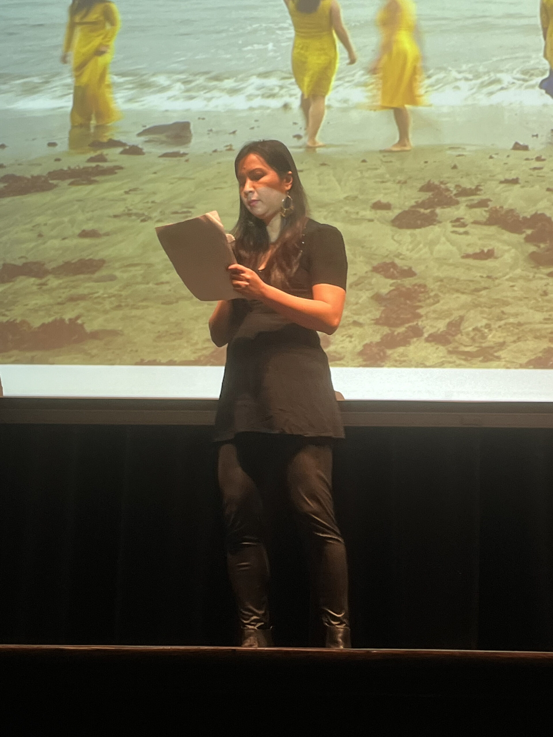 A woman in a black outfit stands on a stage reading from a piece of paper. Behind her is a projection of several women in yellow dresses walking along a beach. The lighting emphasizes her focused expression during what appears to be a live reading or performance.