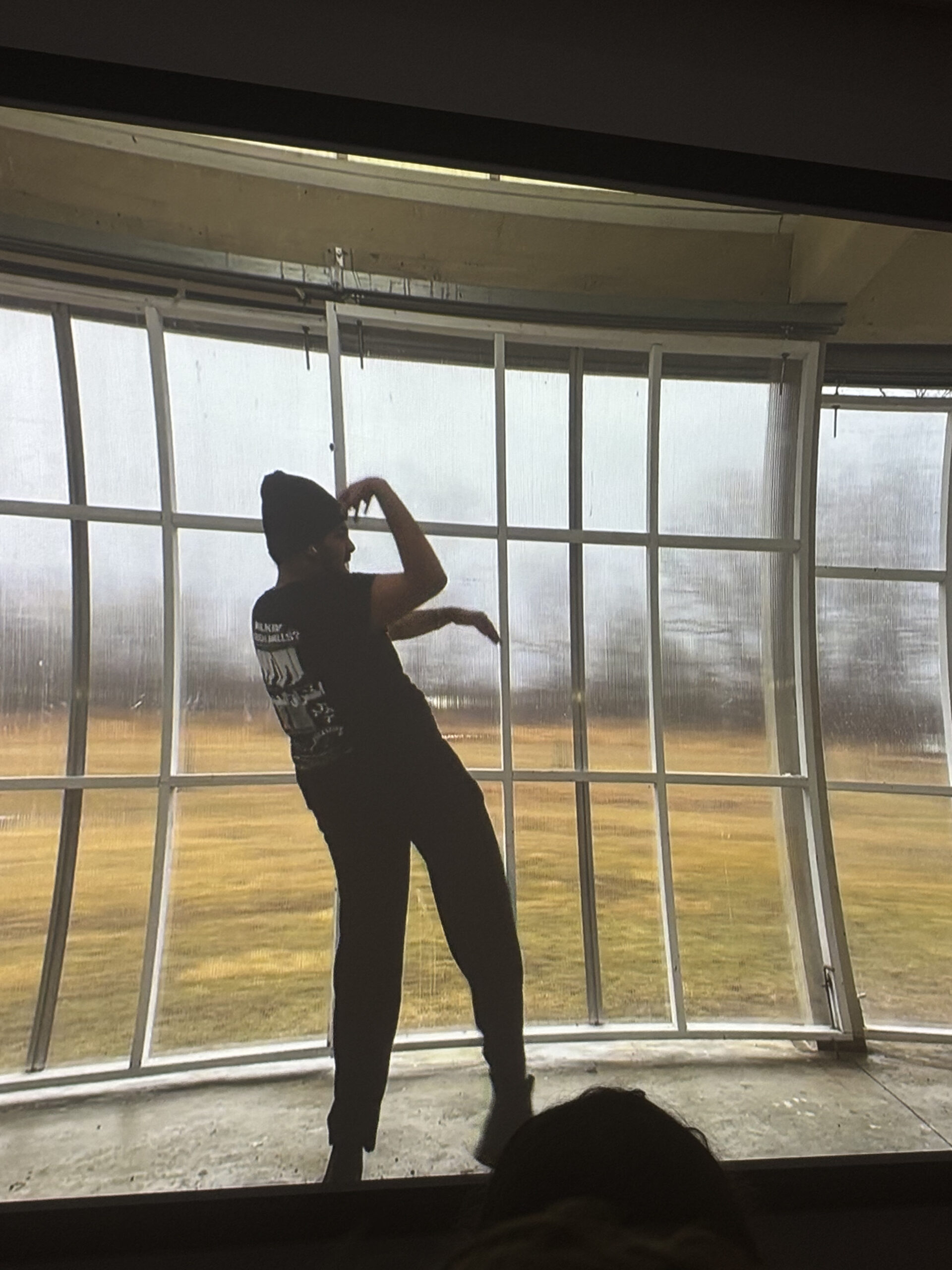 A person dressed in black dances in front of a large, curved window on a rainy day. The wide glass panes show a grassy landscape outside. The figure strikes a dramatic pose, mid-motion, with their arm raised and bent.