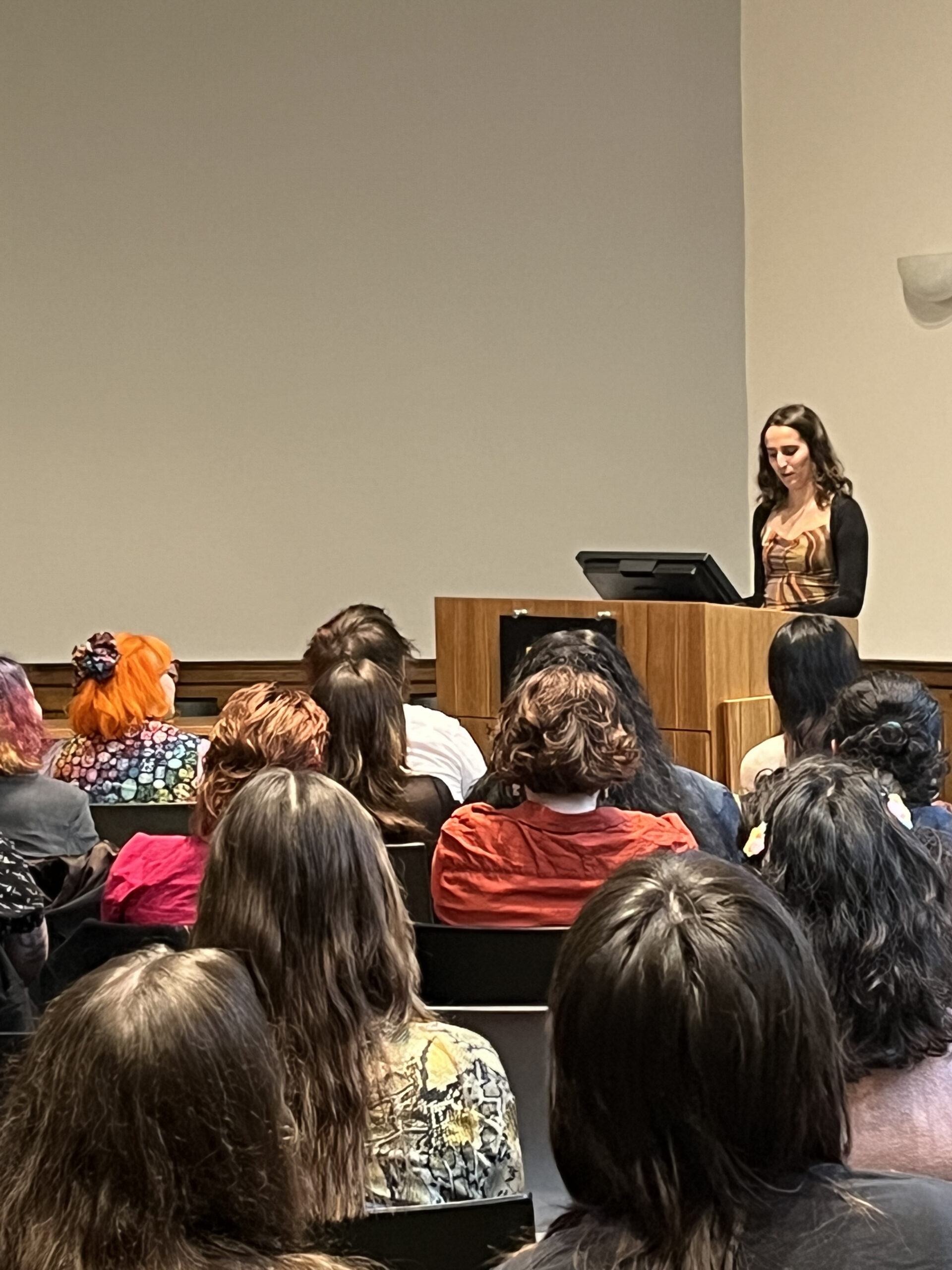 A woman in a brown dress with a black cardigan stands at a wooden podium speaking to an audience. The room is filled with seated attendees, many with colorful hairstyles, watching her intently.
