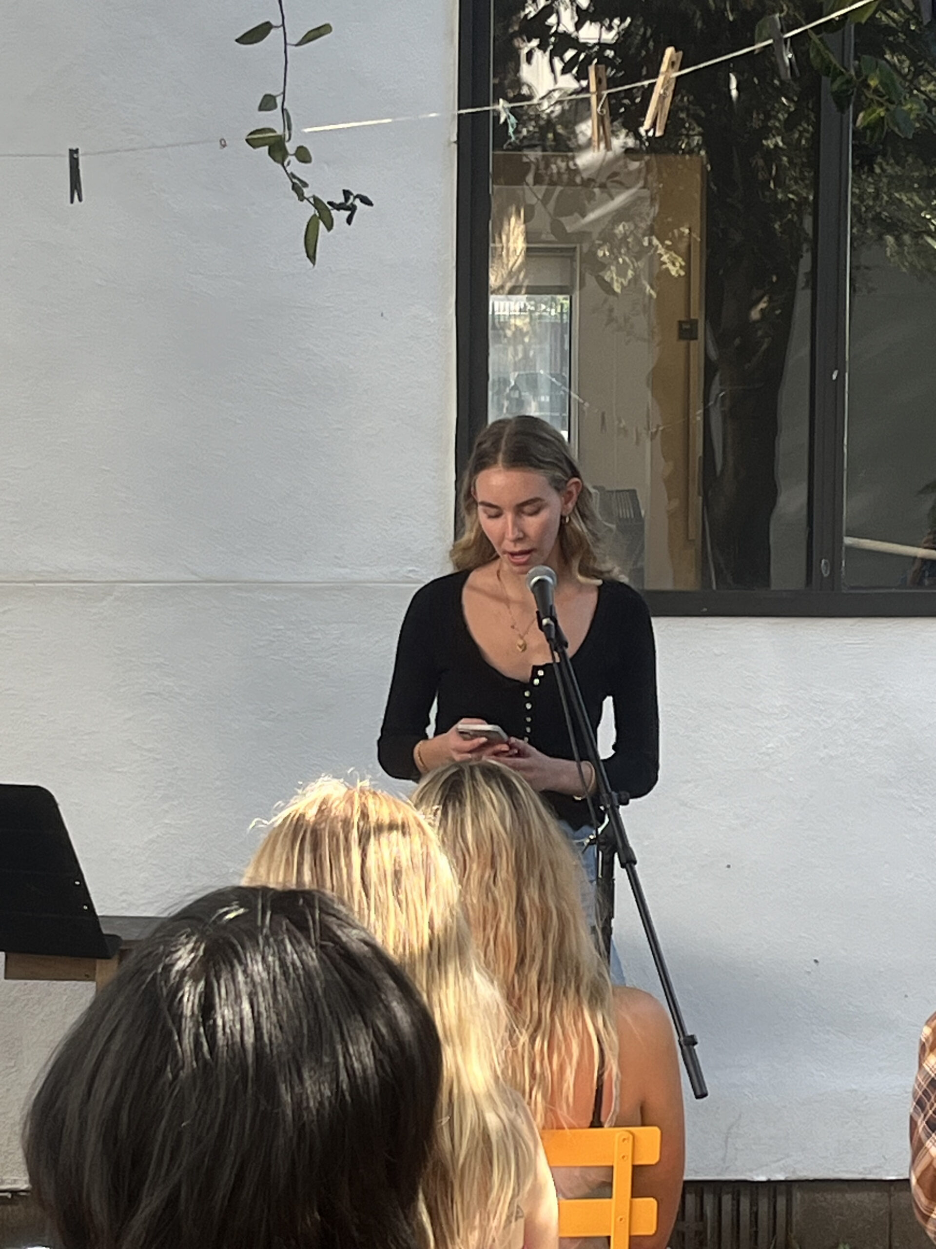 A young woman stands outdoors in front of a microphone, reading from her phone. She is dressed in a black top and jeans. The audience, seated on yellow chairs, listens attentively in a garden setting with string and clothespins overhead.