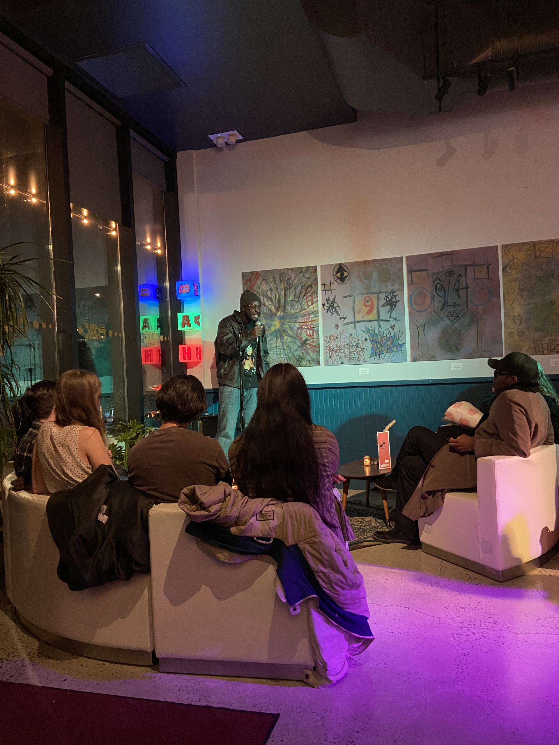 A man in a beanie and leather jacket performs stand-up comedy with a microphone in a cozy venue. The audience, seated on white couches, watches attentively. Behind him are colorful abstract paintings on the wall and glowing letter blocks in a window.