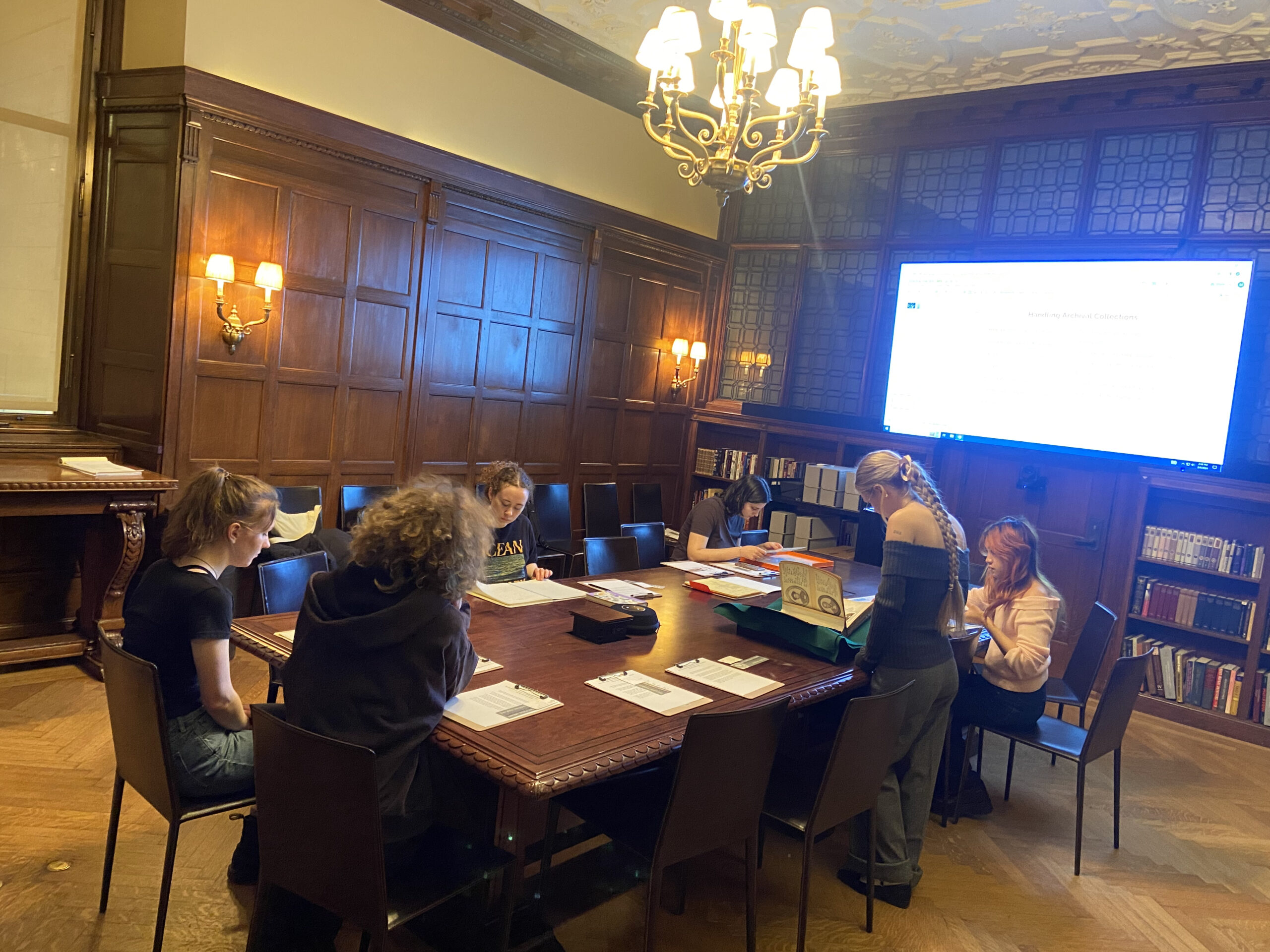 A group of students sits and stands around a large wooden table in a historic library room with ornate paneling and a chandelier overhead. They are engaged in archival research or a class activity, with books, papers, and folders spread across the table. A large monitor on the wall displays a document labeled “Humbling Archival Collections.” Shelves of books and archival boxes line the room.