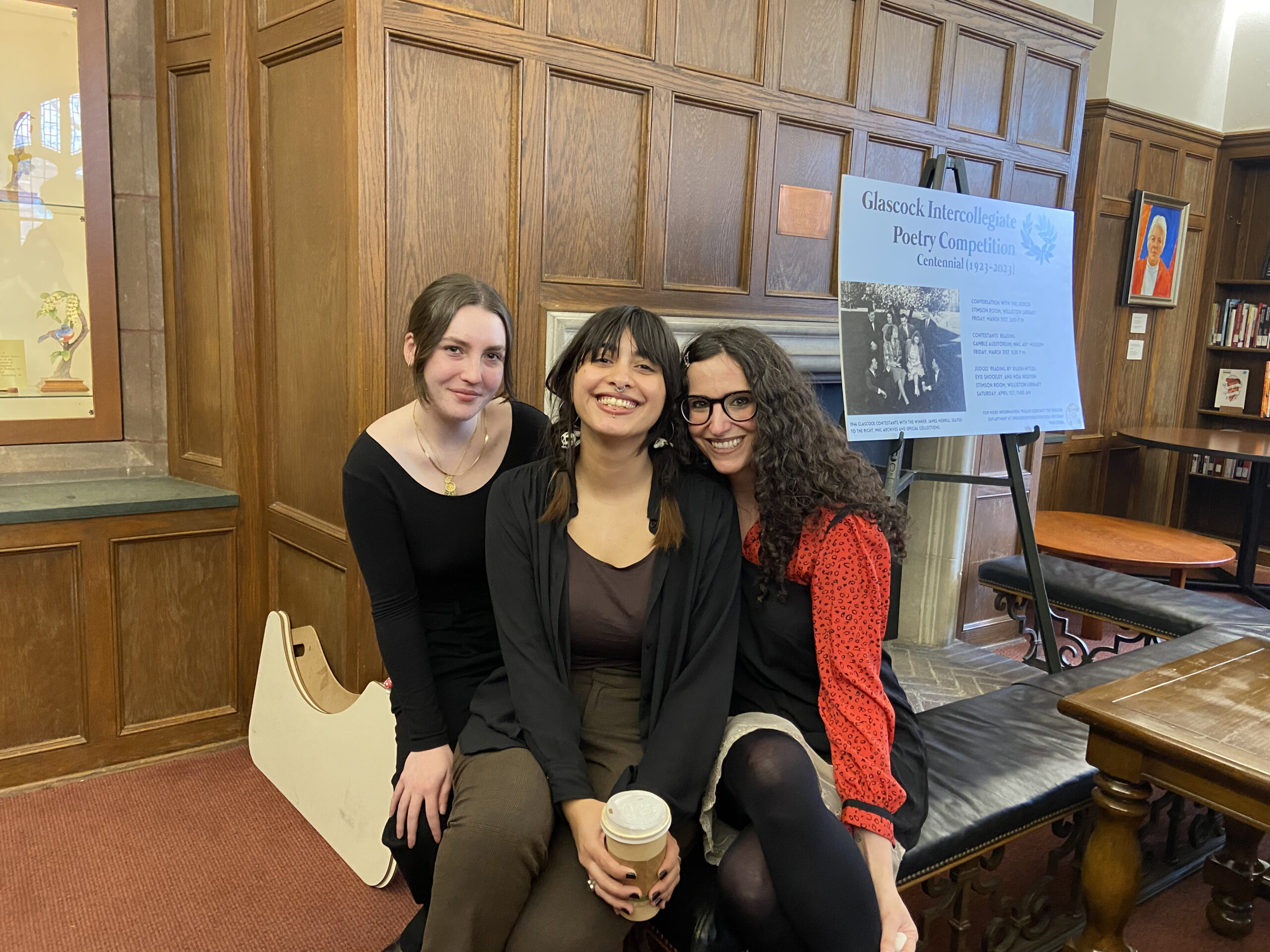 Three women sit closely together inside a wood-paneled library or study room, smiling warmly at the camera. A sign in the background reads “Glascock Intercollegiate Poetry Competition Centennial (1923–2023),” indicating a literary or academic setting.