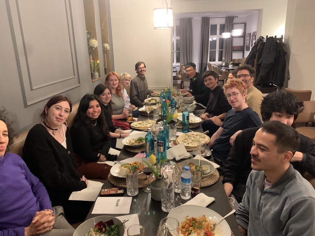 A group of approximately 16 students and faculty sit around a long dinner table in a warmly lit restaurant. The table is set with plates of food, glasses, and blue glass bottles, while the attendees smile and engage with one another across the table.