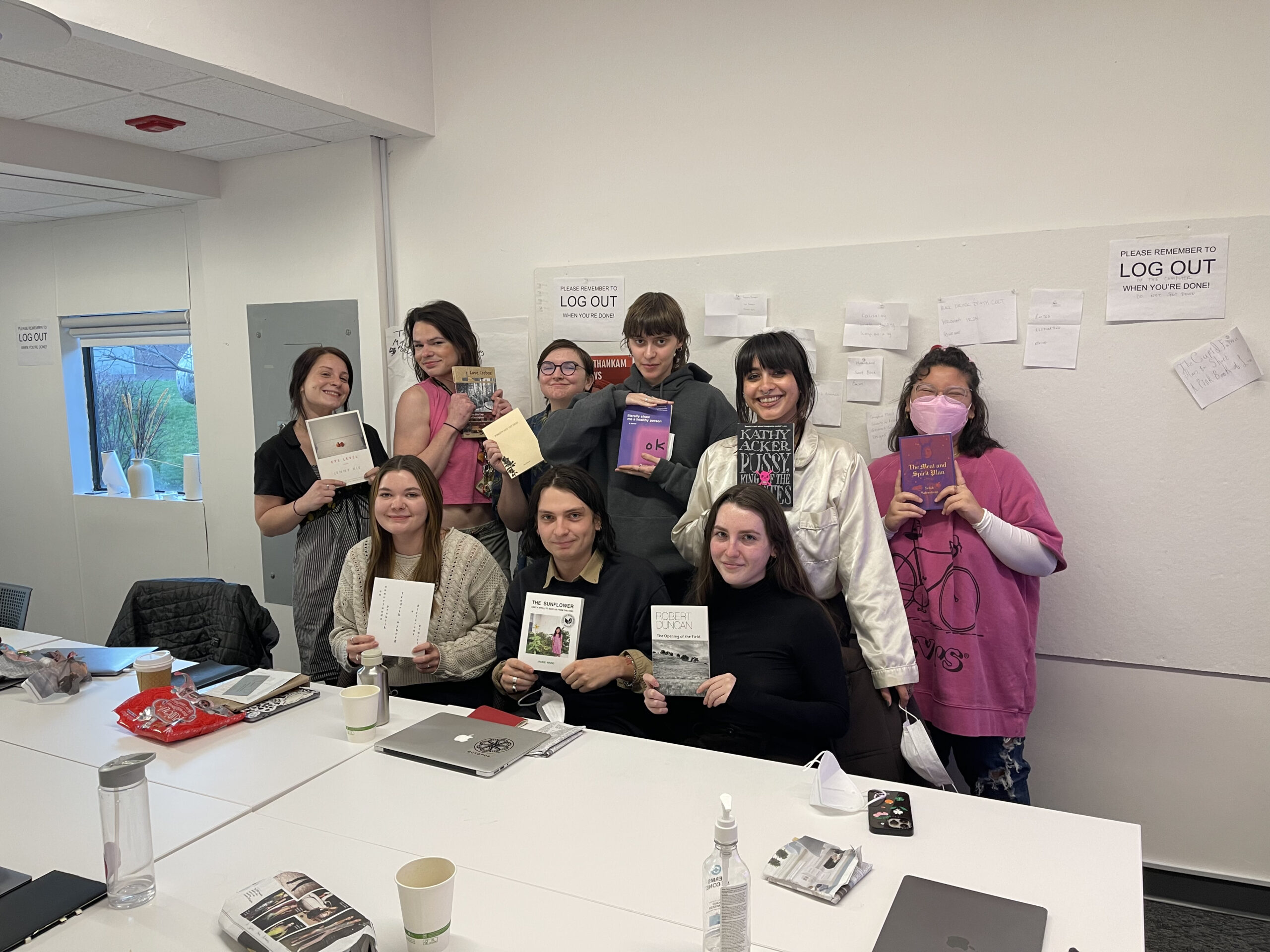 Ten people, mostly students, pose indoors in a classroom holding up various books, showcasing them proudly. They stand and sit behind a white table cluttered with coffee cups, laptops, and papers. The atmosphere is informal and cheerful, with posters and signs on the wall reading “LOG OUT” behind them.