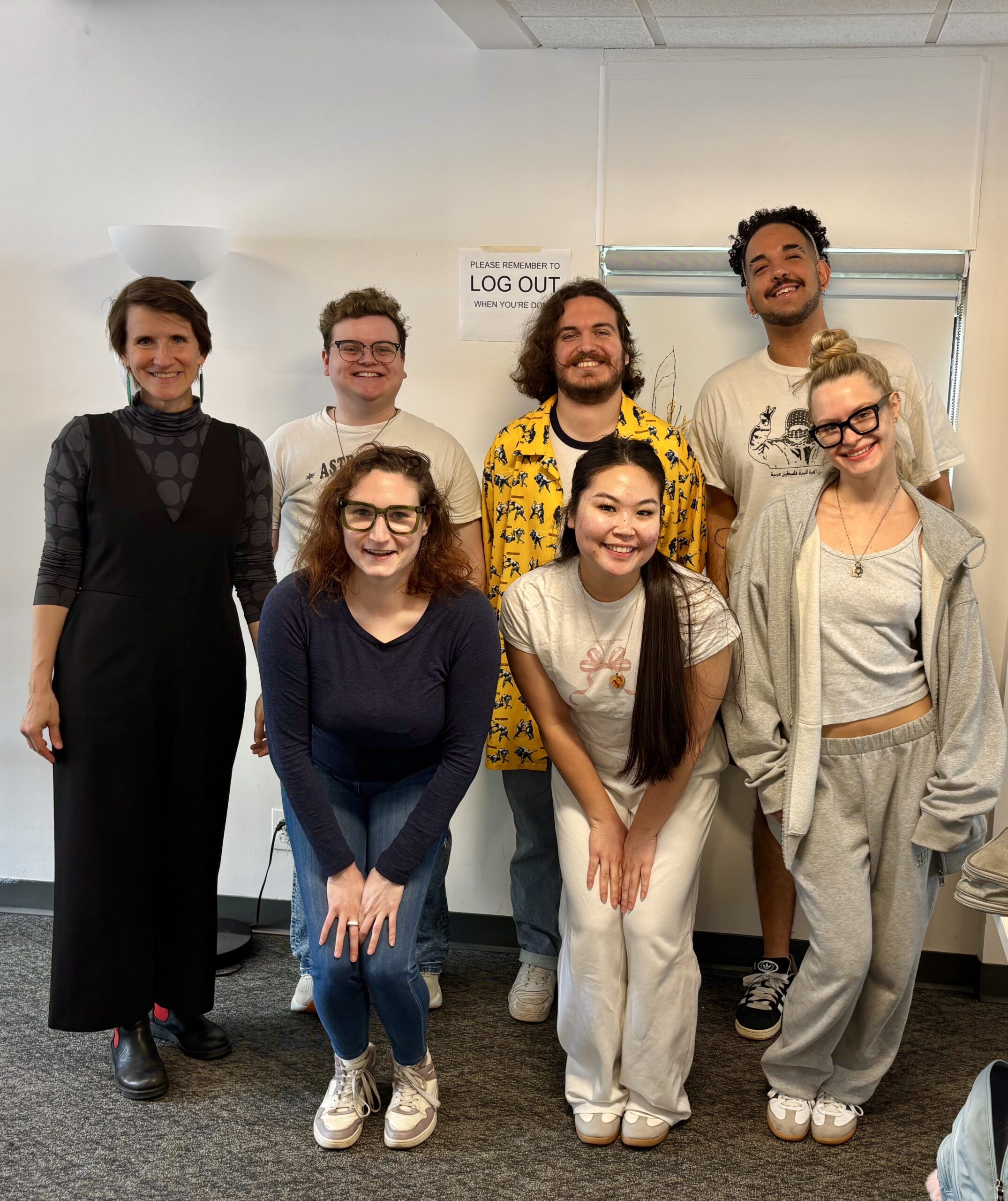 A smiling group of eight students and their professor pose in a classroom in front of a white wall. Everyone is casually dressed, with the professor on the left wearing a black dress over a polka-dot top. A sign behind them reads “Please Remember to Log Out.”