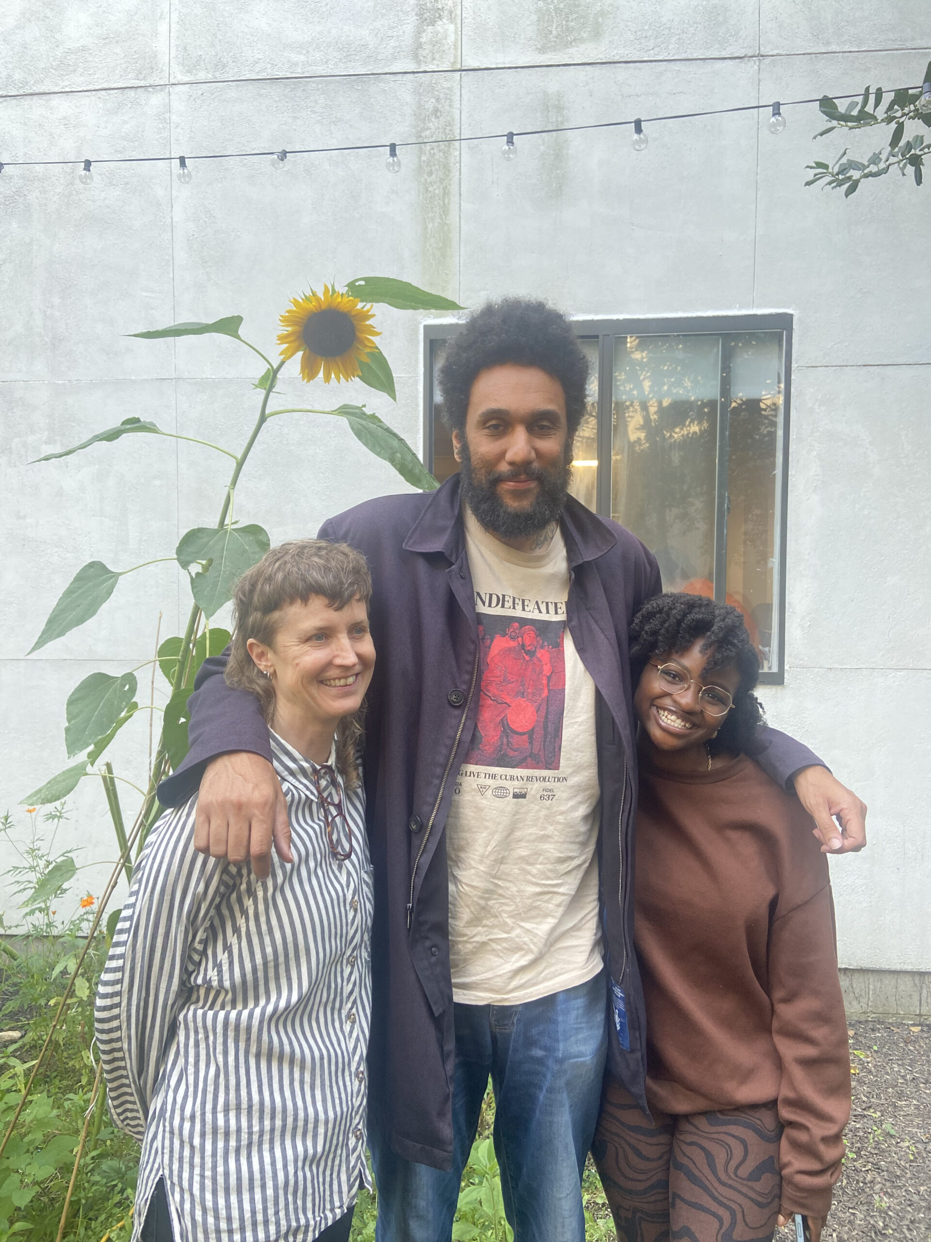 Three people stand smiling in an outdoor garden next to a tall sunflower. The person in the middle has their arms around the shoulders of the other two. Behind them is a modern concrete building with a window and string lights. The mood is relaxed and joyful.