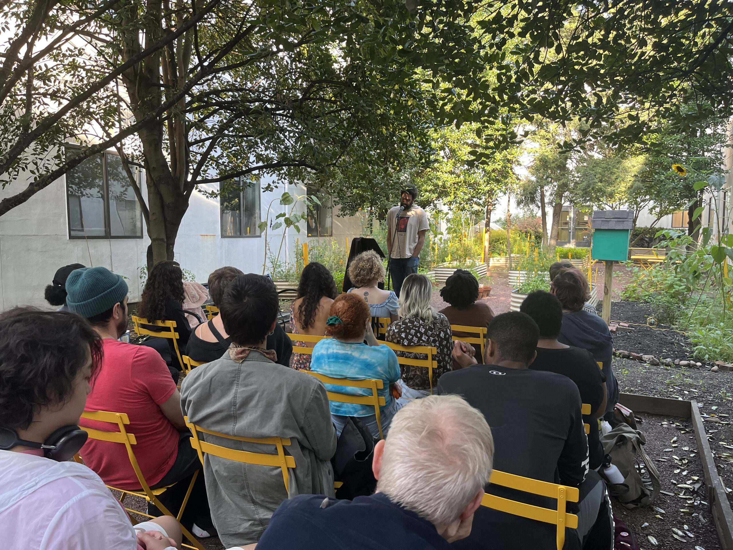An outdoor poetry reading is taking place in a garden courtyard surrounded by trees and greenery. The audience, seated on bright yellow chairs, faces a man standing at the front with a microphone. The man appears to be reading or performing, with sunlight filtering through the trees above. Raised garden beds and a green birdhouse add to the community garden atmosphere.