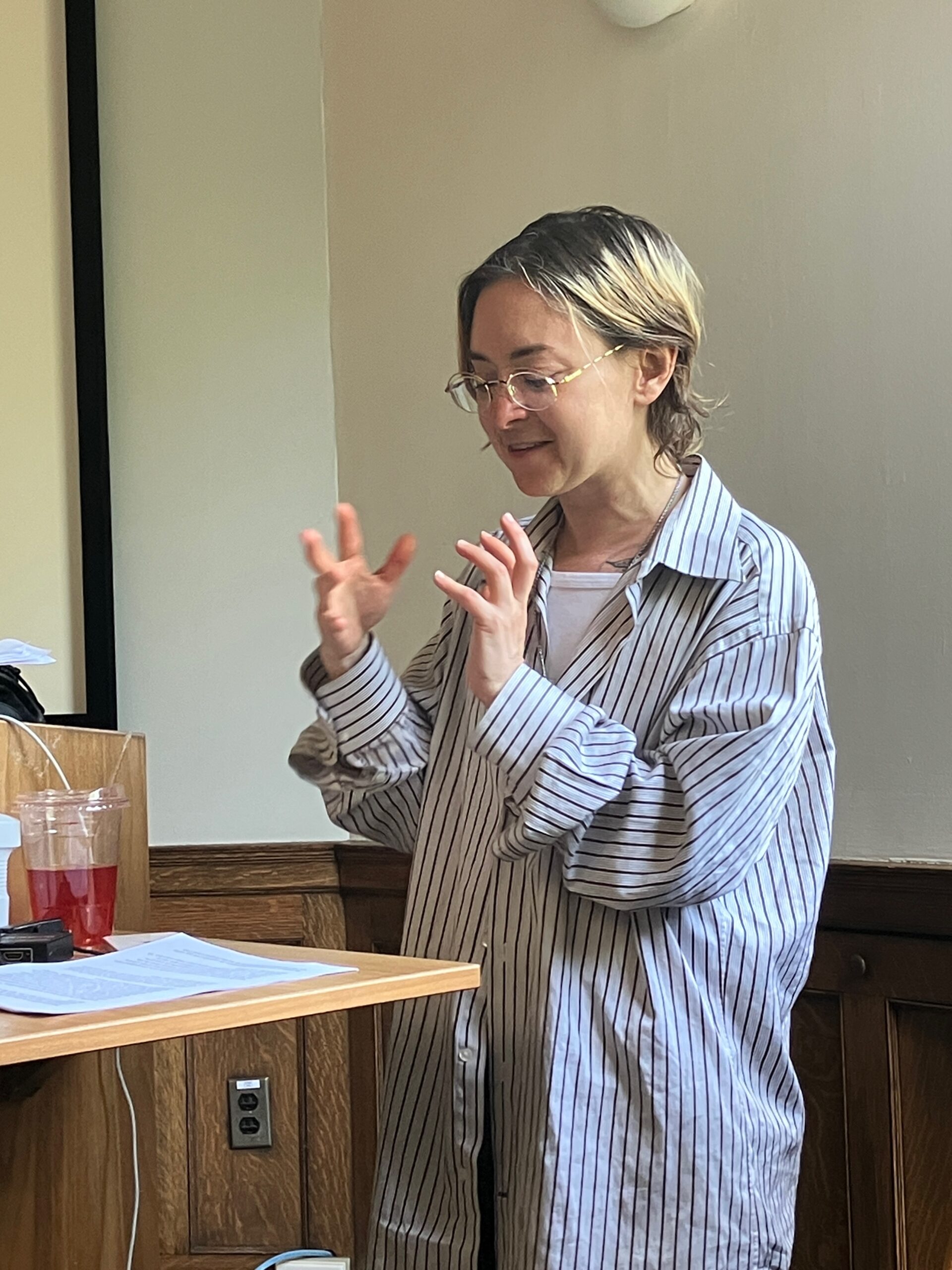 A person wearing glasses and a loose striped shirt gestures expressively while speaking at a lectern. Papers, a drink with red liquid, and electronic equipment are on the small desk in front of them. The setting is a classroom with wood paneling and natural light.