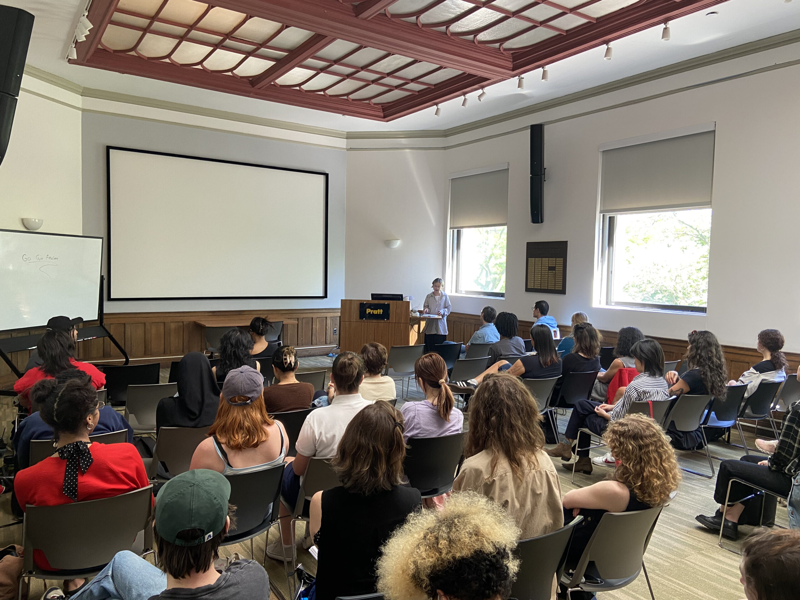 A speaker stands at a podium in a large classroom or lecture hall with rows of students seated and facing forward. Behind the speaker is a large blank projection screen and a whiteboard with the words “Go Go Future” written on it. The atmosphere is formal and academic, likely a guest lecture or seminar.