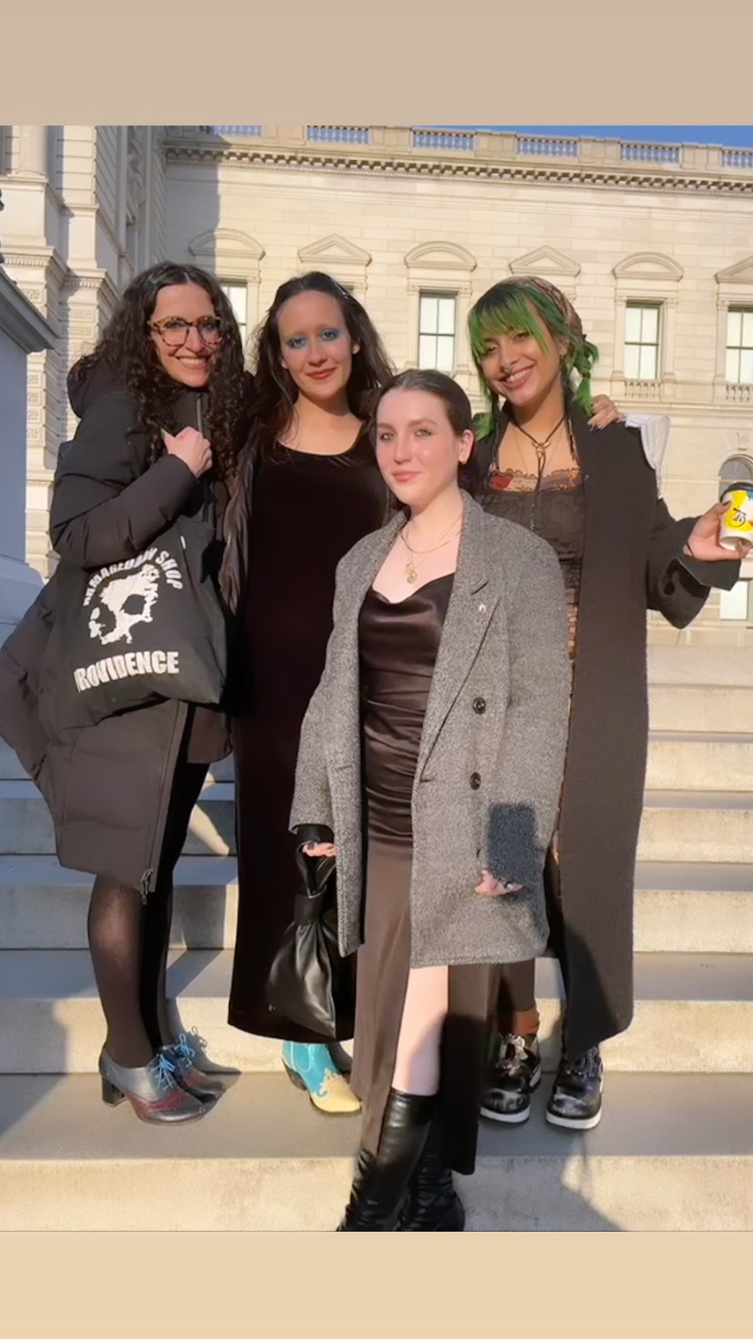 Four individuals stand together on stone steps in front of a neoclassical government building, smiling and posing in afternoon sunlight. Each person wears a dress or coat, and the individual on the far left carries a black tote that reads