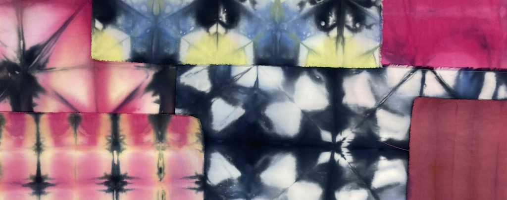 A patchwork textile composition featuring tie-dye and dyed fabric squares in shades of pink, blue, yellow, black, and white. The fabrics display symmetrical, kaleidoscopic patterns and gradients, overlapping to form a layered abstract design.