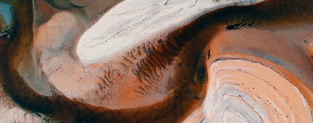 Aerial view of a natural landscape with curving, layered earth formations in shades of rust orange, brown, white, and gray. The textures suggest eroded sediment or mineral deposits, resembling abstract patterns from above.