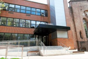 Exterior view of Pratt Institute’s North Hall building showing a modern red-brick façade with large windows, a metal-clad vertical accent panel, and a black canopy above the entrance steps. A ramp with metal railings provides accessible entry to the building.