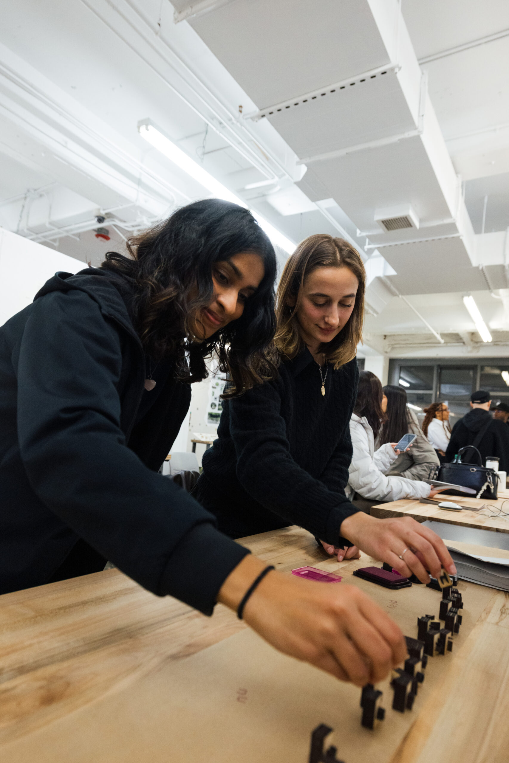 Two women are engaged in a creative project at a long wooden table. One woman has dark, wavy hair and is placing a small block onto the table, while the other, with straight hair, looks on and also interacts with similar items. The scene is set in a well-lit room with people in the background, and various materials are spread across the table.