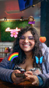 A smiling person with curly hair and glasses sits at a table, holding a smartphone in their hands. They wear a floral top under a puffy jacket. Colorful emojis, including a peach, rainbow, butterfly, flower, and a woman gesturing, are superimposed around them. The background features greenery and a softly illuminated sign.