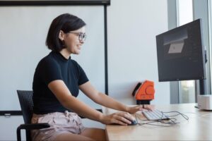 A young woman with short hair, wearing glasses and a black top, smiles while working on a computer at a desk. She uses a mouse and keyboard, with a monitor displaying a design interface. The background features a partially visible orange device and large windows letting in natural light.